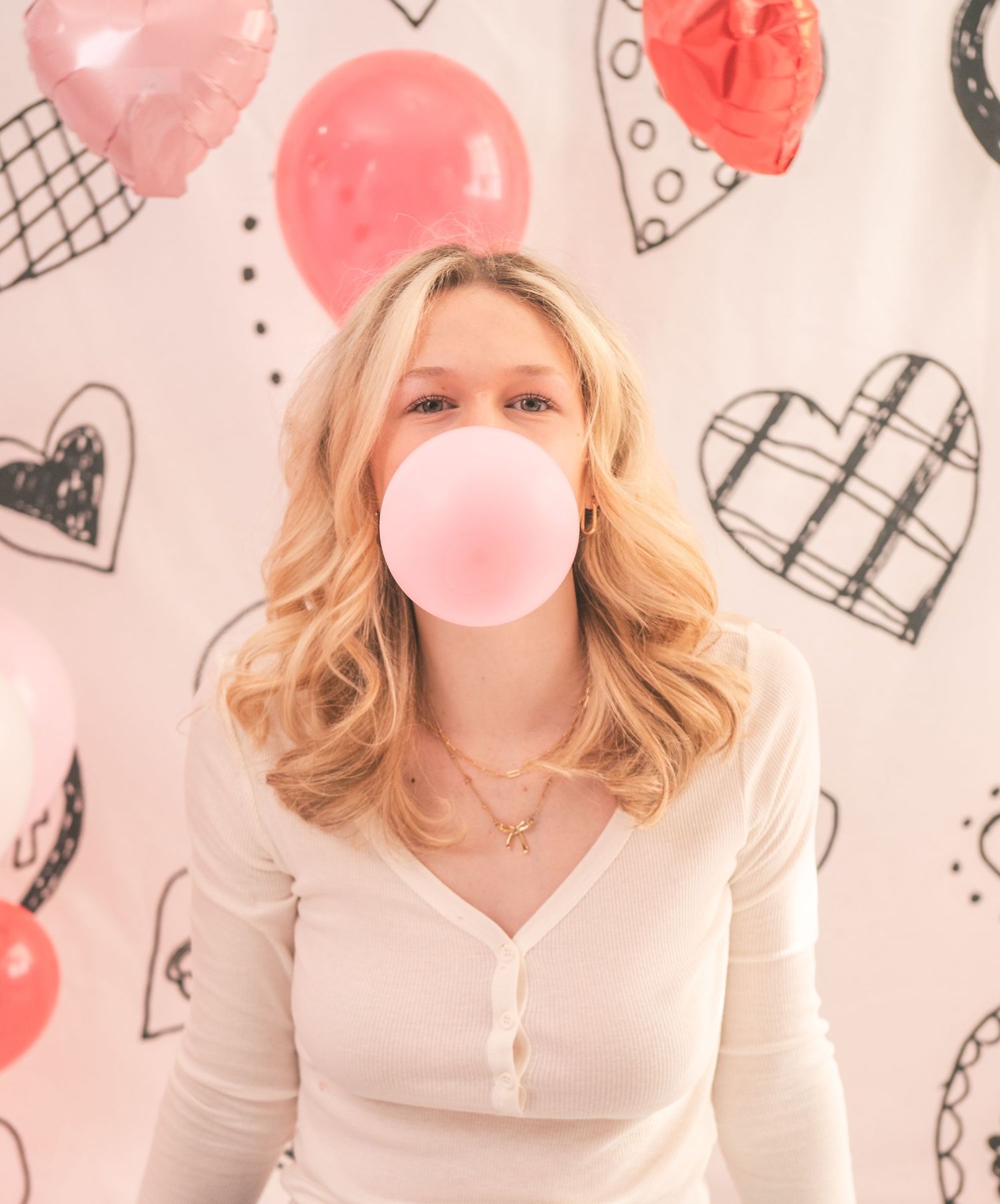 Someone blowing pink bubblegum while posing against a background with pink and black heart pattern doodles.