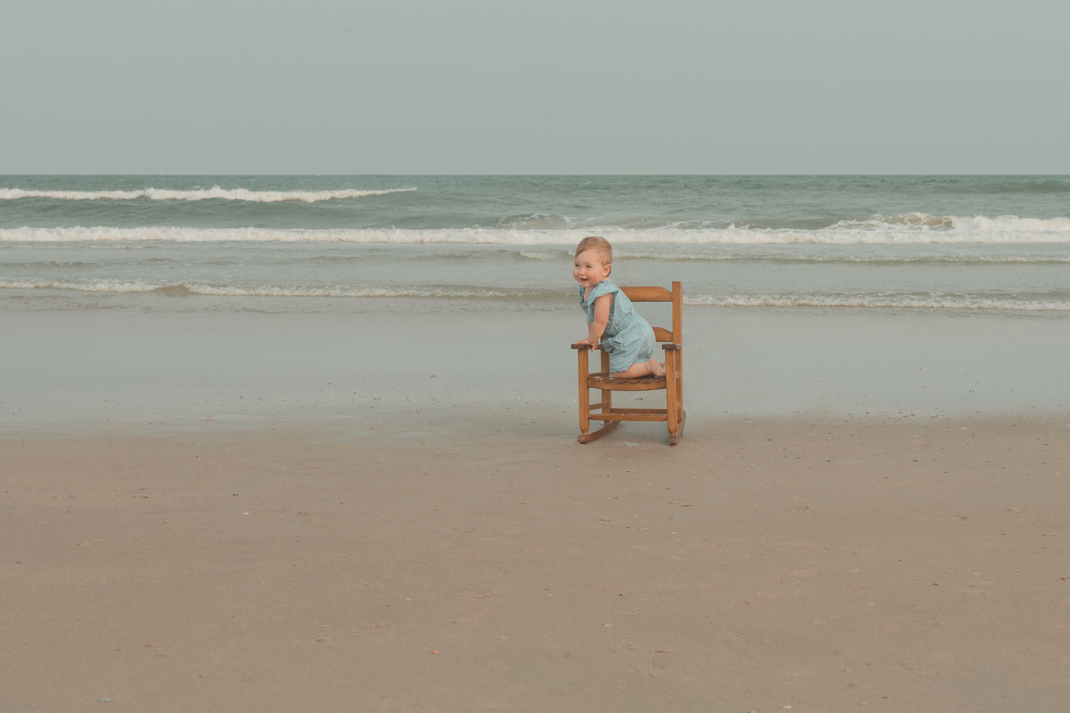 A small wooden rocking chair sits alone on a sandy beach with gentle waves rolling in the distance.