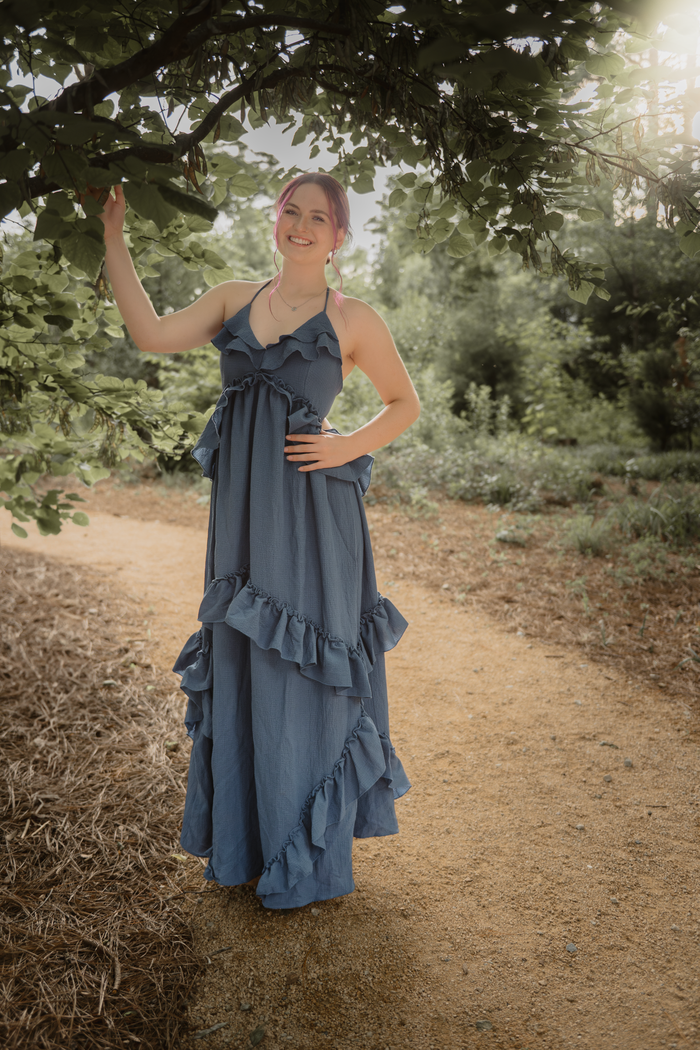 A whimsical portrait of Haylee in a flowing blue ruffled maxi dress standing on a dirt path under green trees during her senior session