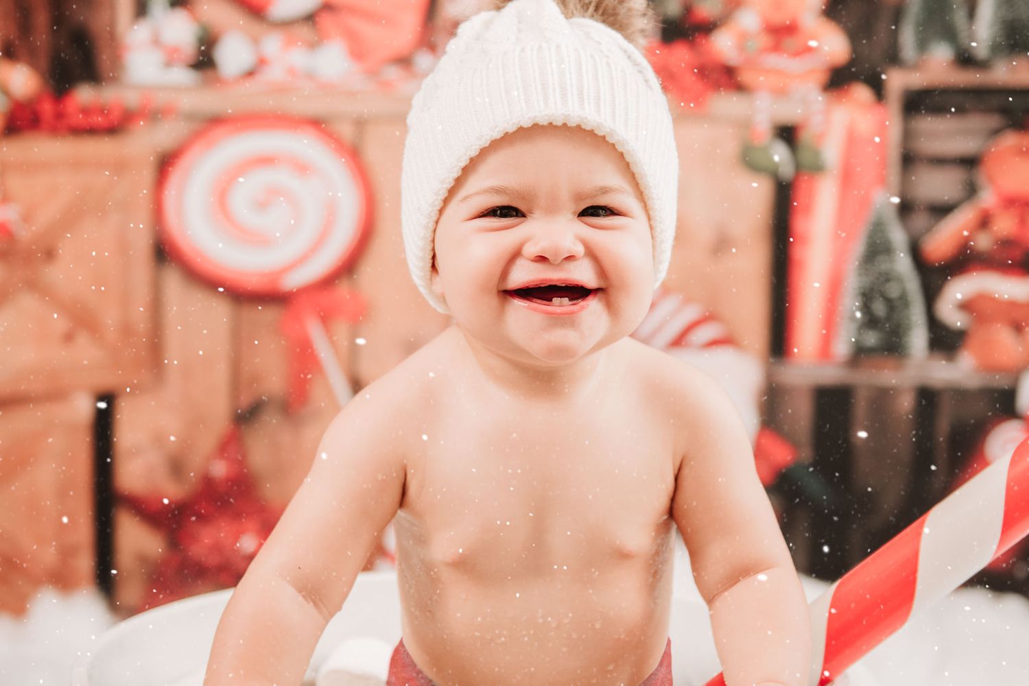 A laughing baby wearing a white knit hat sits among festive red and white Christmas holiday decorations.