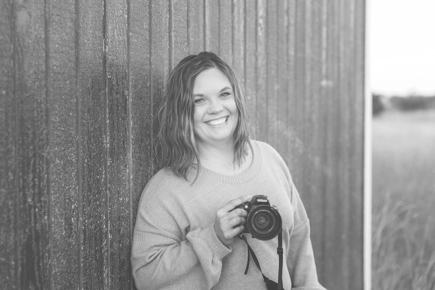 Black and white portrait of smiling photographer holding camera while standing against wooden wall outdoors.