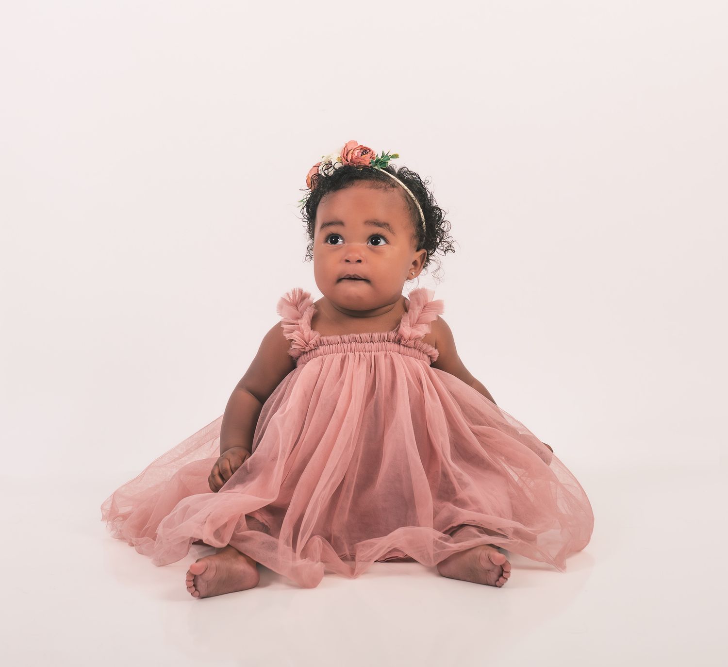 A baby wearing a pink tulle dress and floral headpiece sits on a light studio backdrop.