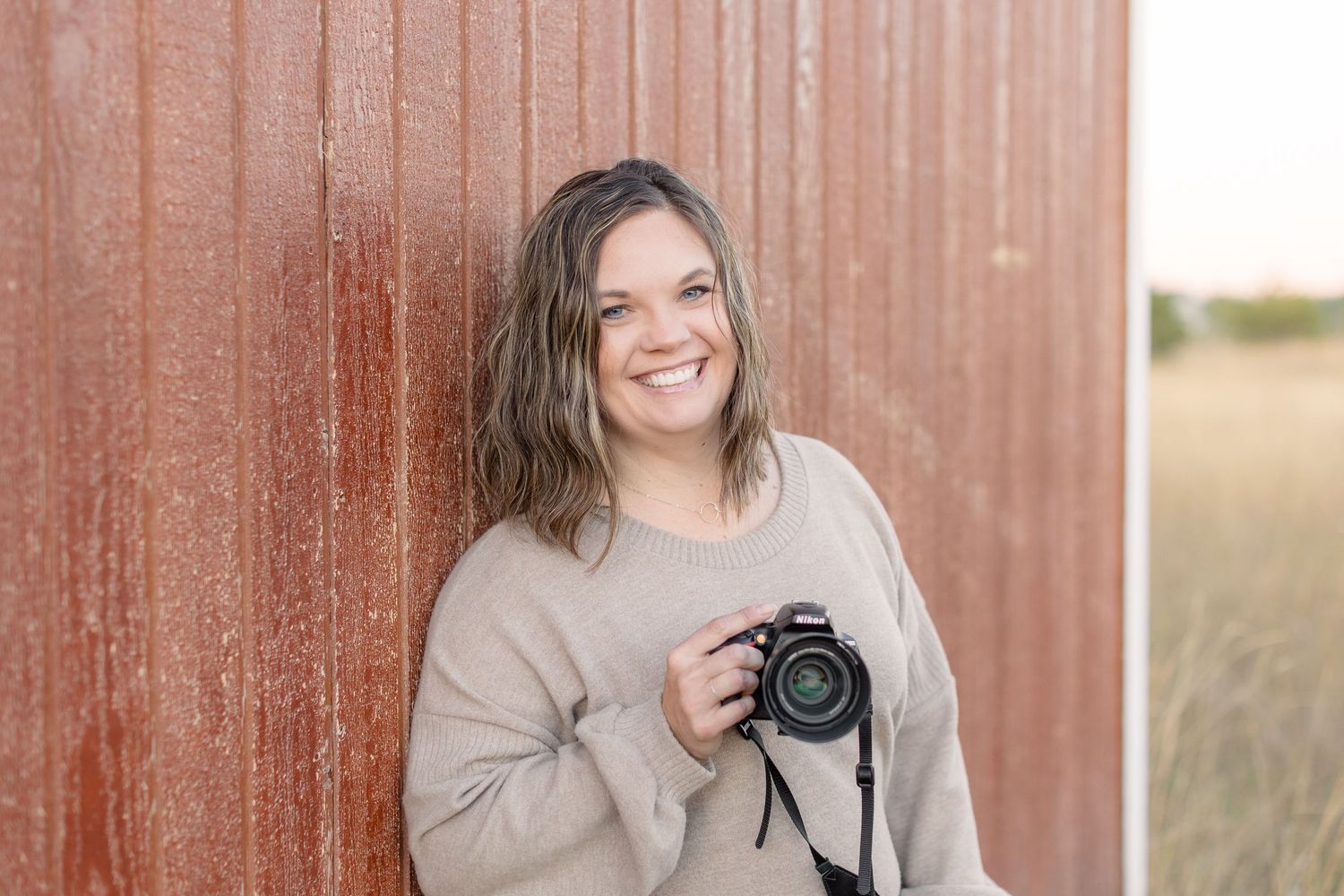 A photographer in a beige sweater holds a professional camera while standing next to a rustic red wall.