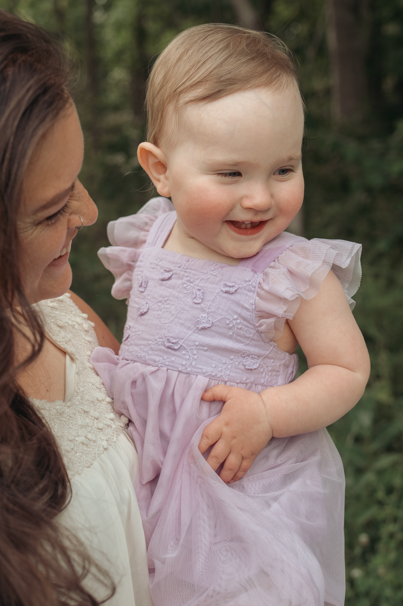 Sweet baby in a pink ruffled dress smiles joyfully while being held outdoors against a green forest backdrop.
