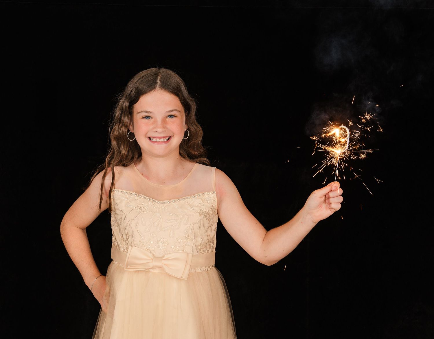 A smiling figure in a cream-colored dress holds a glowing sparkler against a dark background.