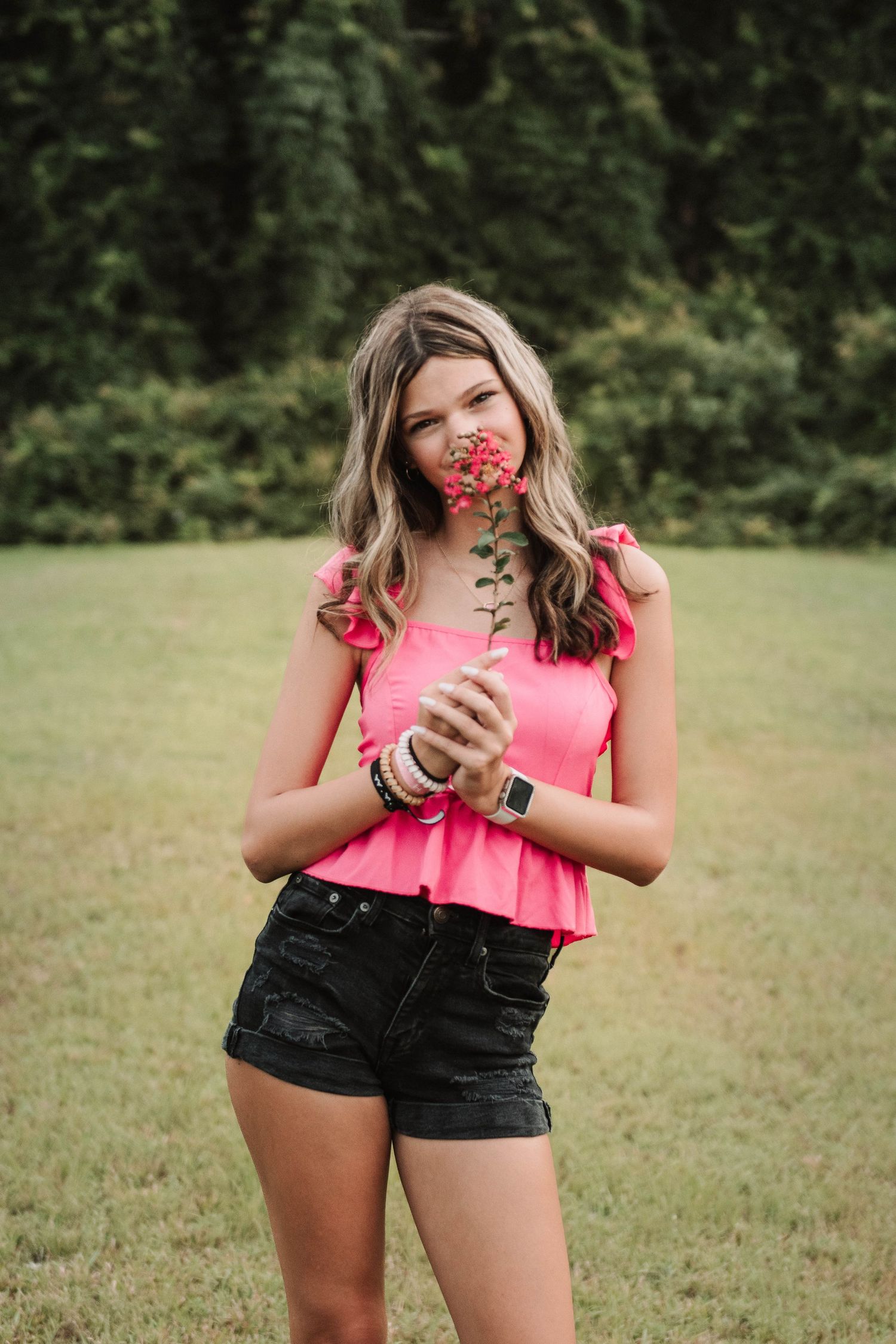 A person in a pink peplum top and dark shorts holds a red flower in a grassy field near woods.