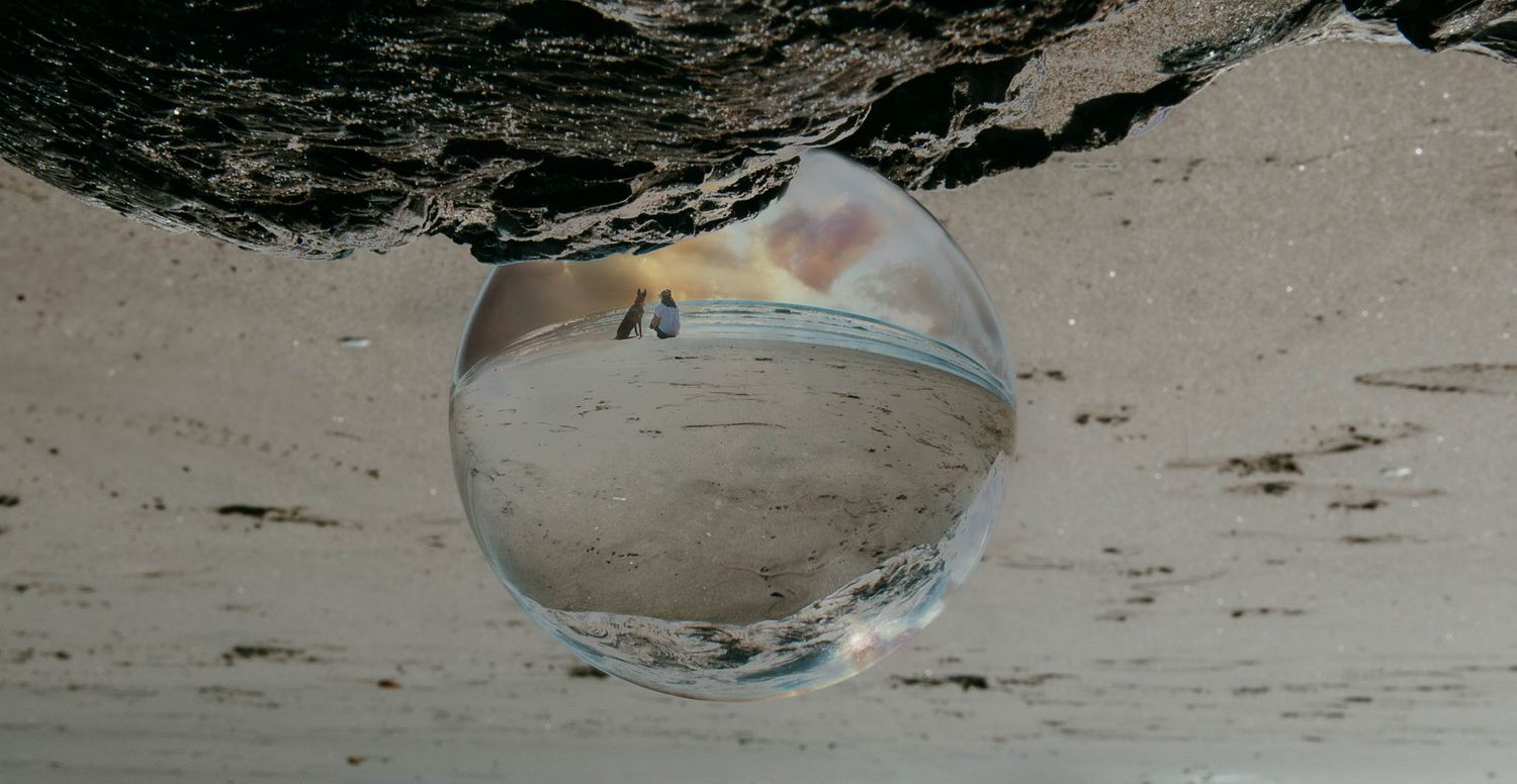 A clear glass orb sphere reflects a beach scene with sand and ocean waves against a blurred background.
