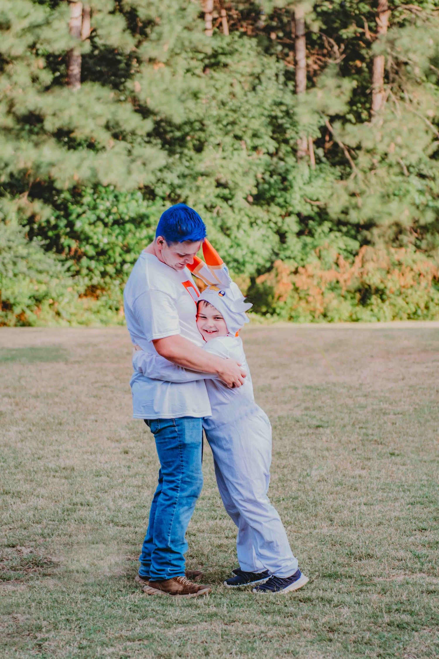 Two people in casual clothing share an embrace while standing in a grassy field with pine trees in the background.
