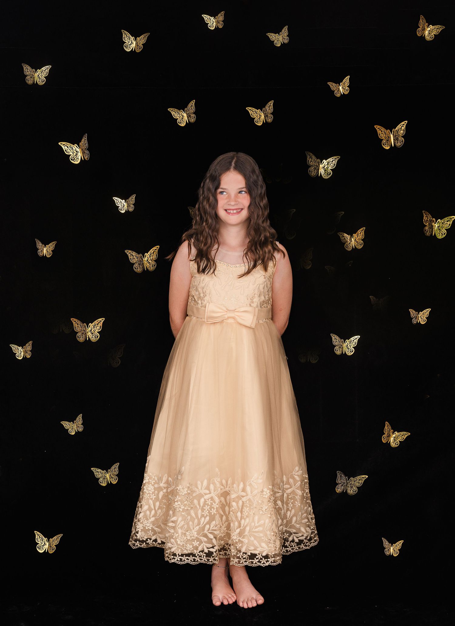 A figure in a cream-colored dress stands barefoot against a black background with golden butterflies floating around.