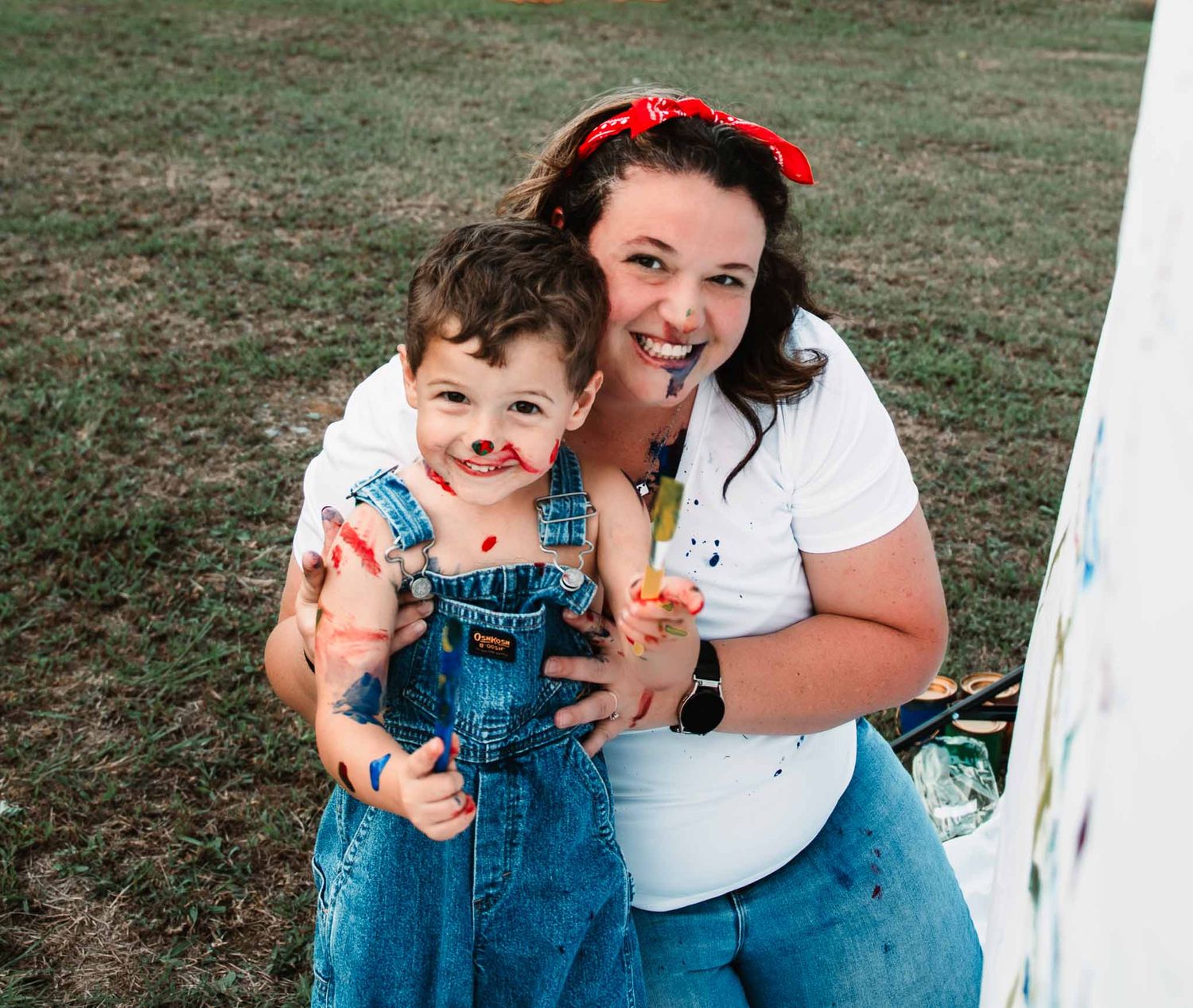 A cheerful scene on grass featuring a person in a white shirt and red headband with a child wearing denim overalls.