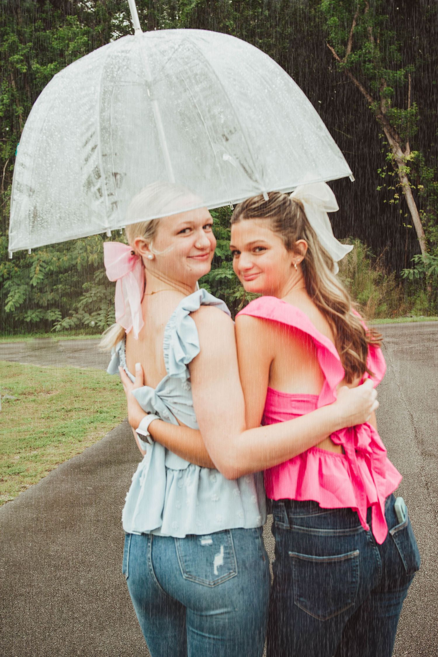 Two friends sharing a clear umbrella while wearing ruffled tops and jeans on a rainy day.
