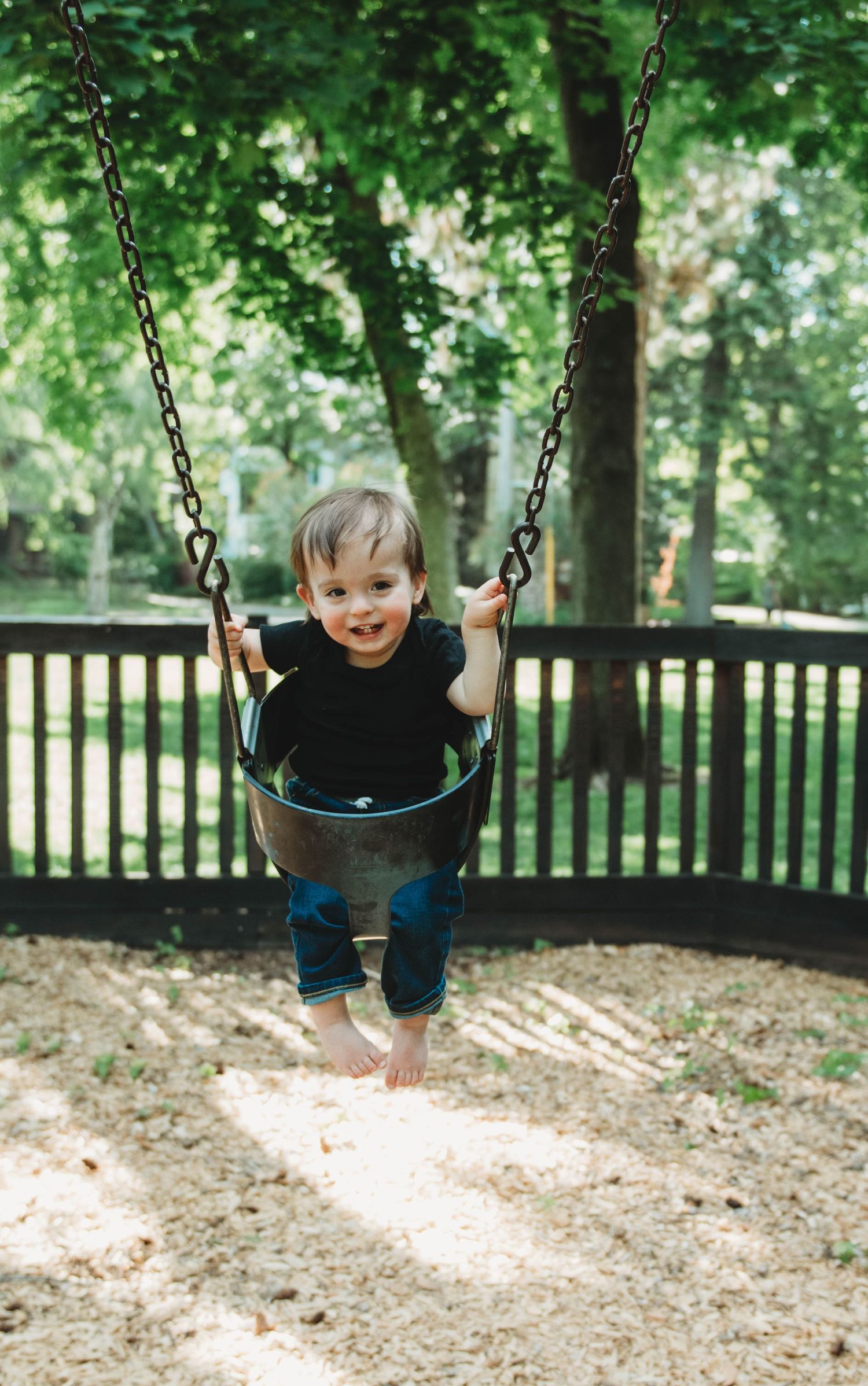 A child in a black shirt swings on a playground swing set surrounded by green trees and a wooden fence.