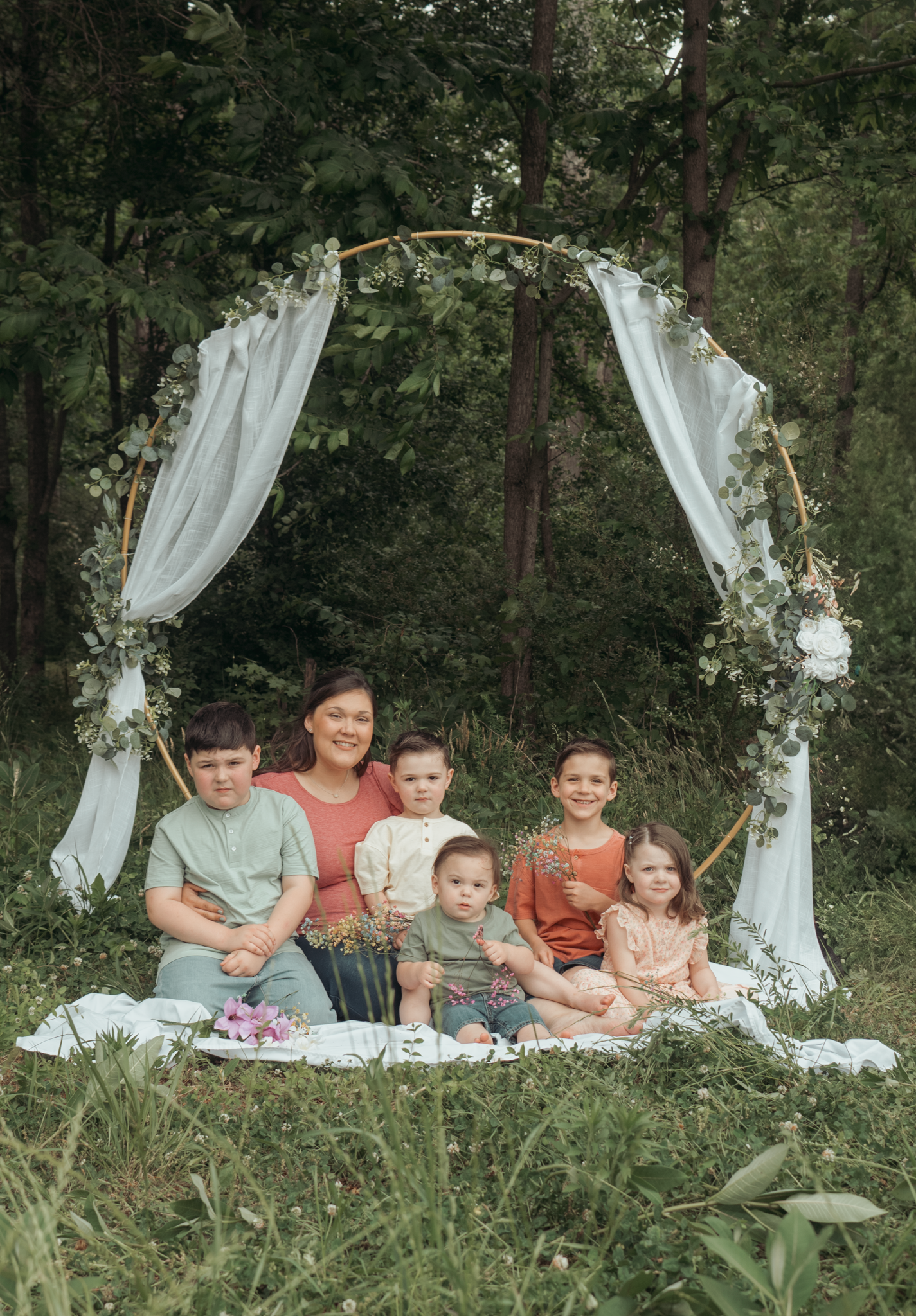 A family group sits on a white blanket under a decorated floral arch with flowing white fabric in a wooded setting.
