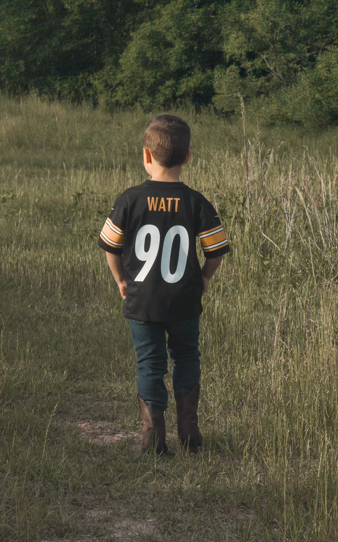 A young Steelers fan wearing a Watt #90 jersey stands in a field looking towards wooded area.