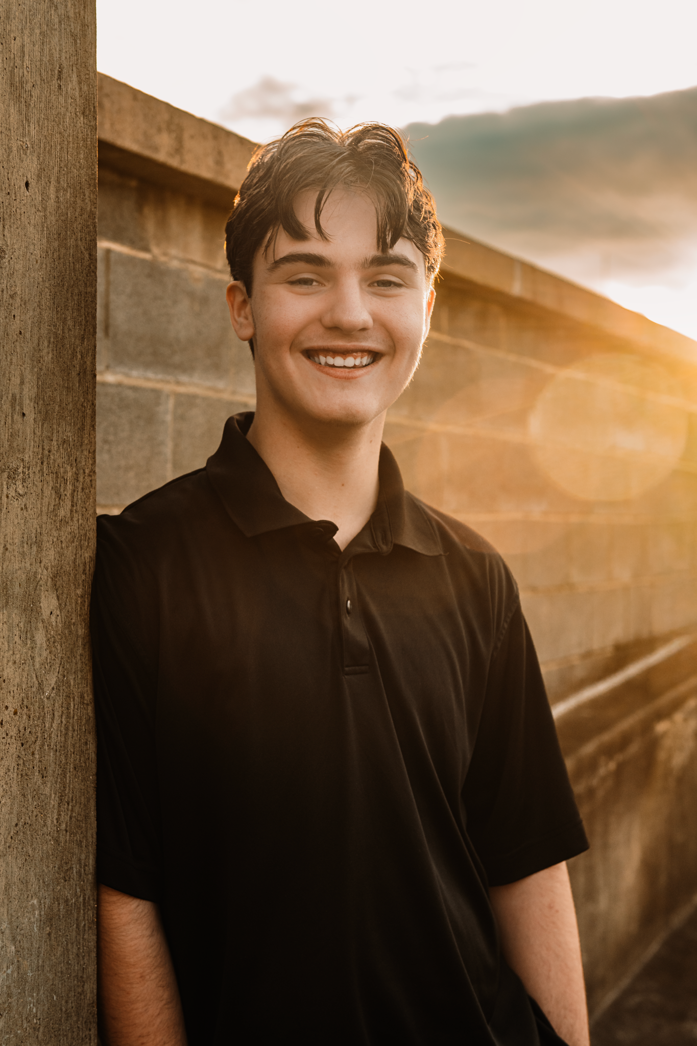 Smiling teenage boy in black polo shirt leaning against stone wall with golden sunset backlight.