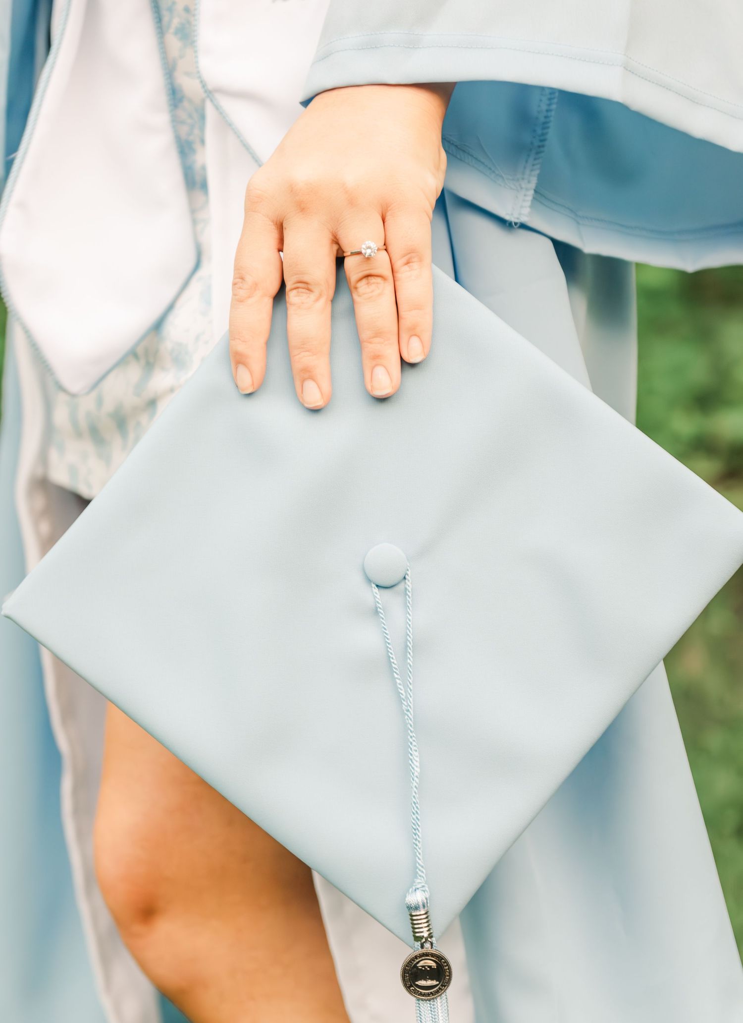 Light blue graduation cap shown with a hand wearing an engagement ring against a soft green background.