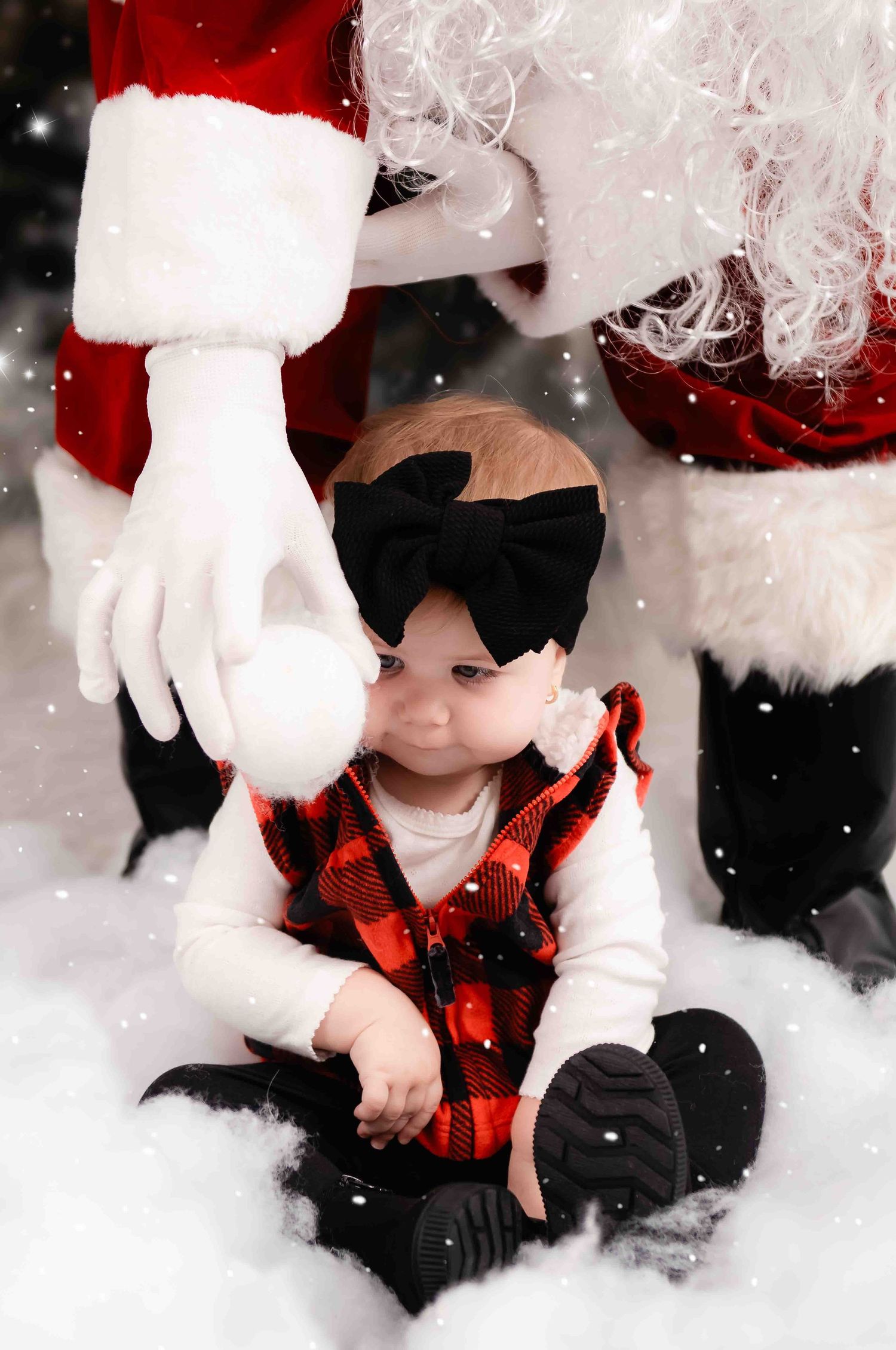 A small child in a black velvet bow and buffalo plaid vest sits in snow near red and white Santa suit details.