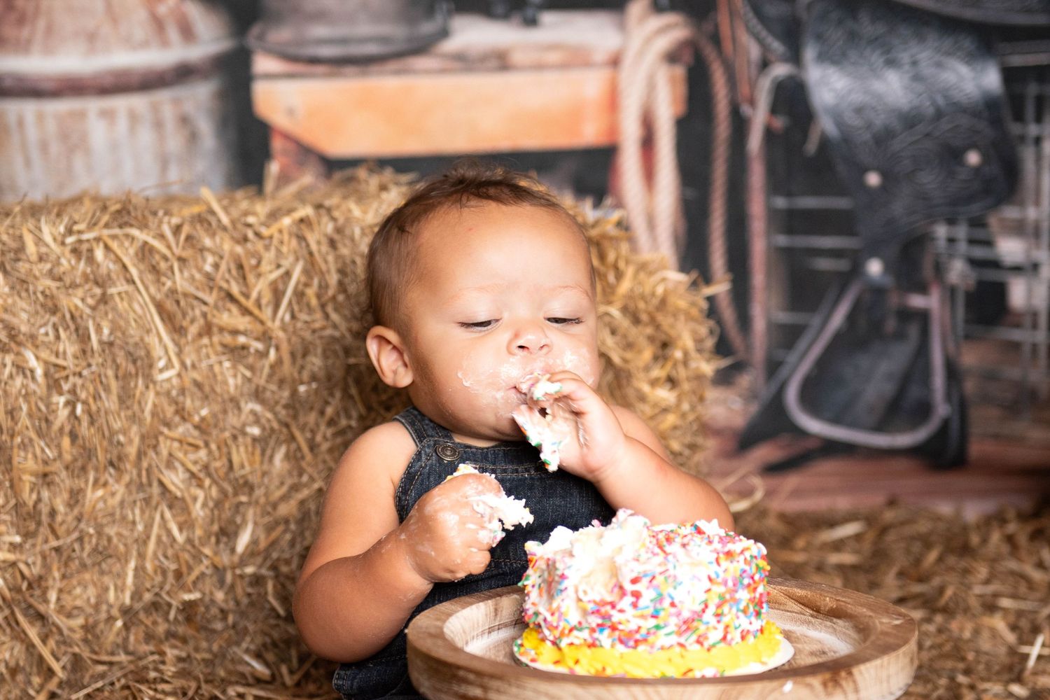 A young child eating cake with white frosting and sprinkles while seated on hay bales in a rustic setting taken by Krystie Nicole Photo