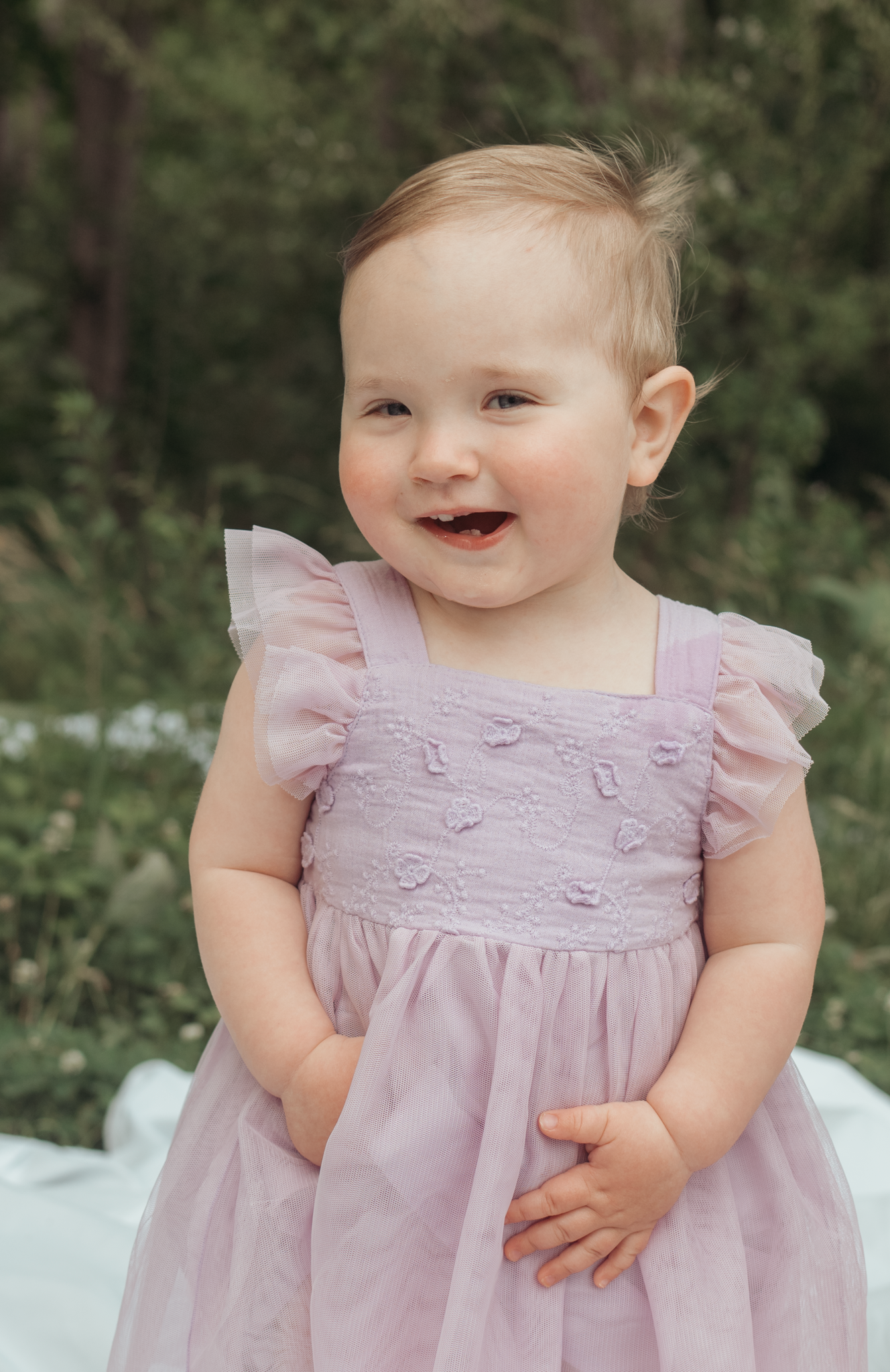 A smiling baby wearing a light pink ruffled dress with embroidered details sits outdoors in a garden setting.