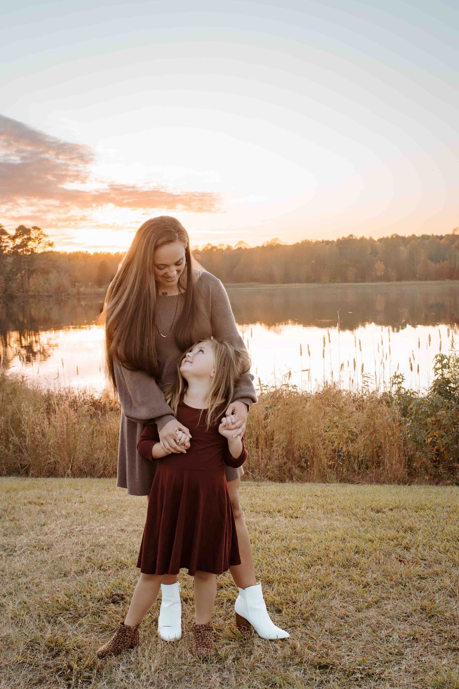 A mom & her daughter, a sweet bonding moment captured during golden hour by a lake with autumn trees and glowing sunset in the background.