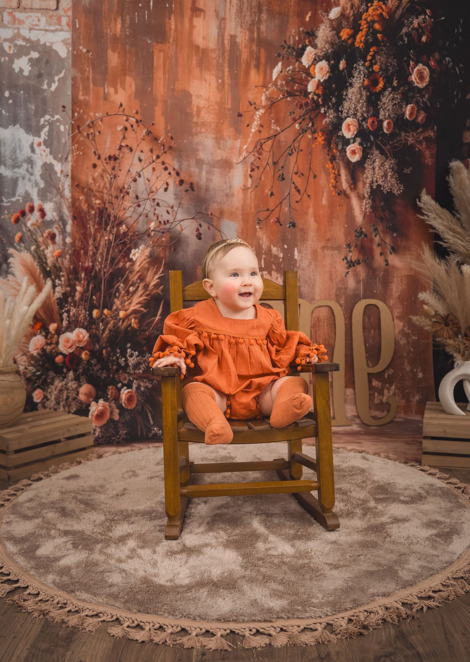 Small child in bright orange outfit sits in wooden rocking chair surrounded by fall floral decorations.