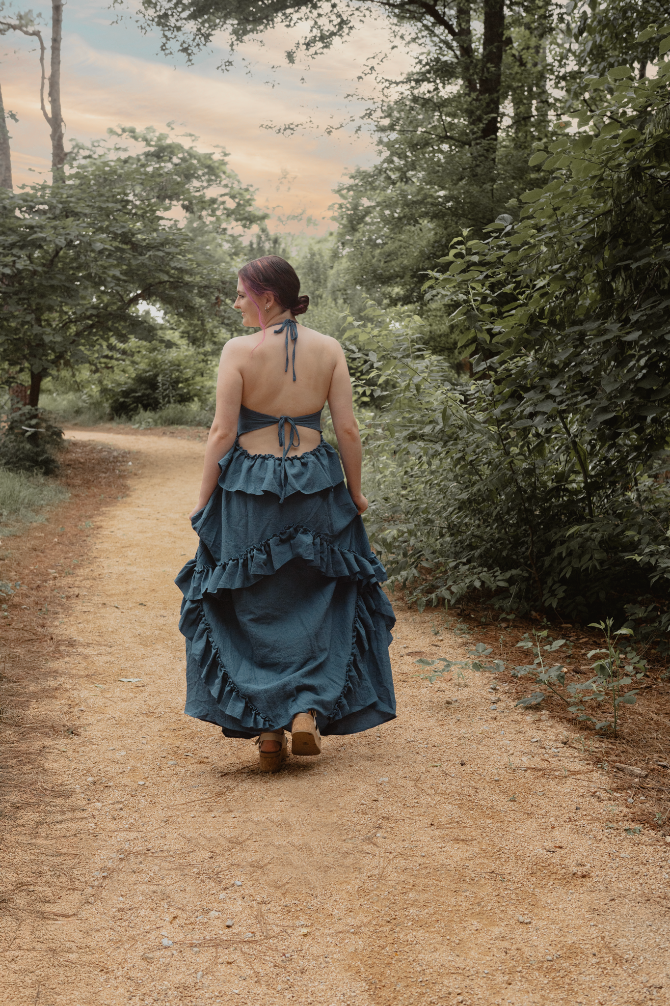 Haylee in a flowing blue ruffled dress walks along a serene forest path during golden hour during her senior session in Raleigh, NC