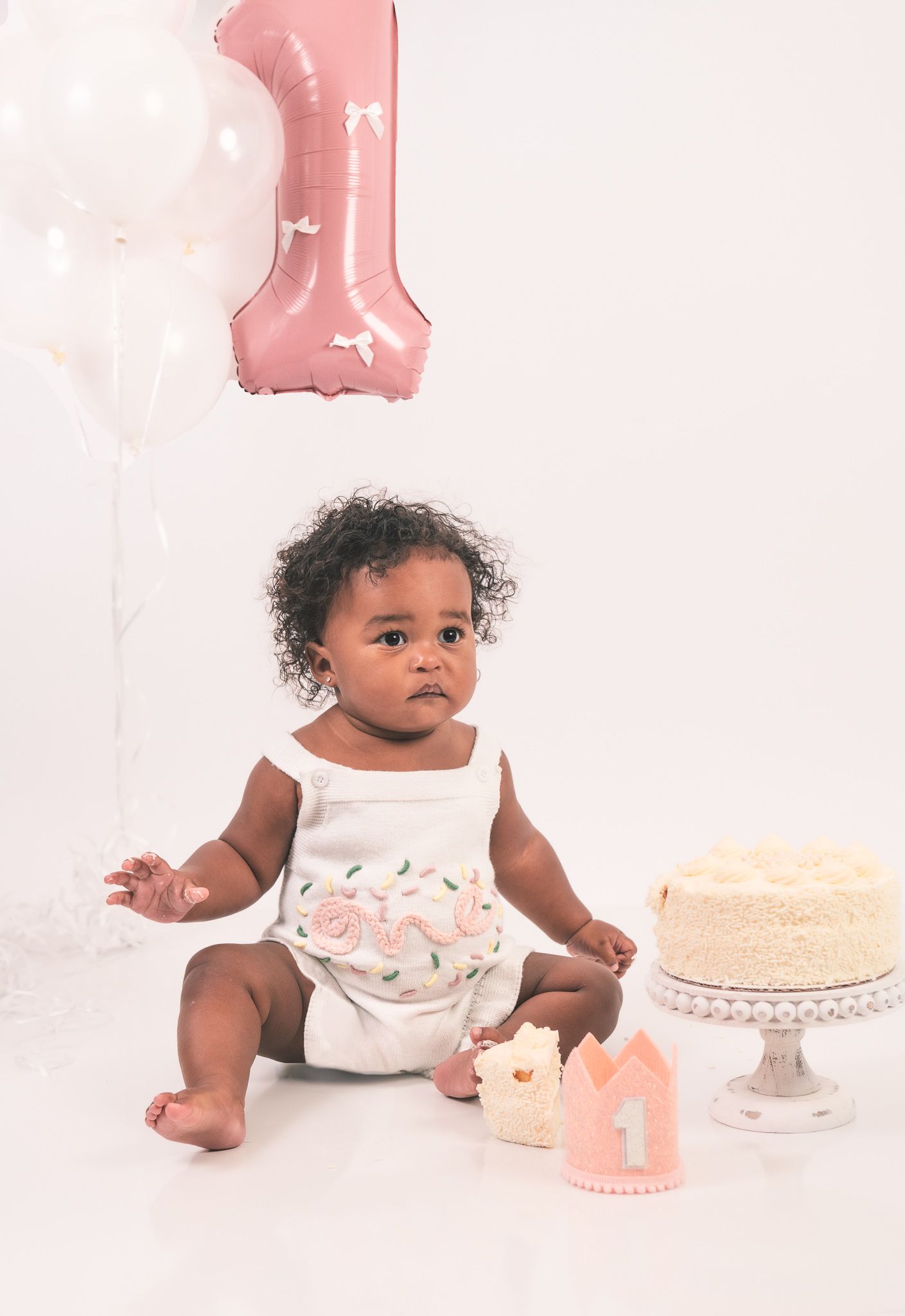 A pink and white first birthday celebration setup with a cake on a stand and number one balloon decoration.