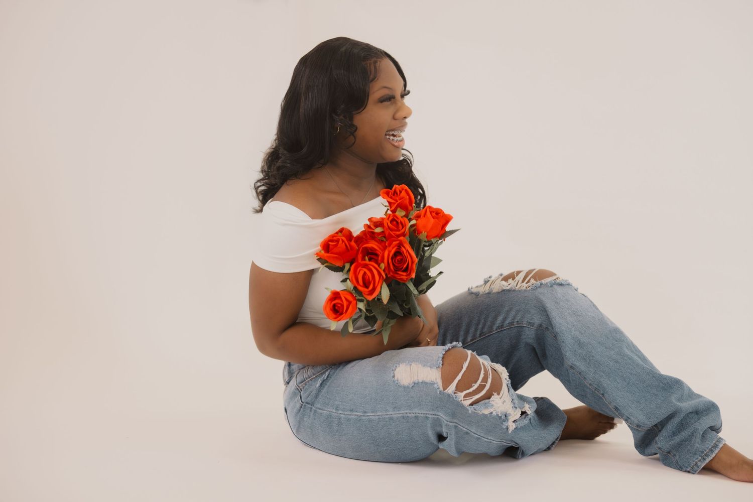 Woman in white off-shoulder top and distressed jeans holds vibrant red poppies while sitting on light backdrop.