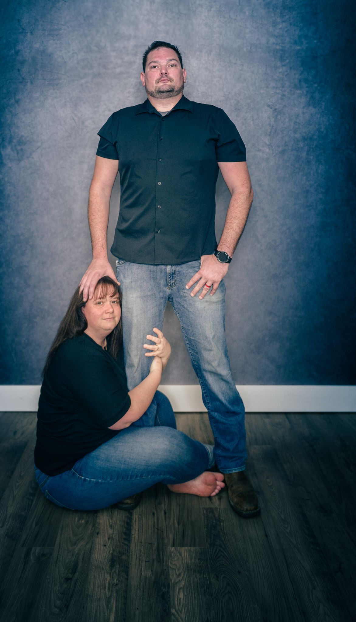 A couple posing for a studio portrait against a blue textured background on hardwood flooring.