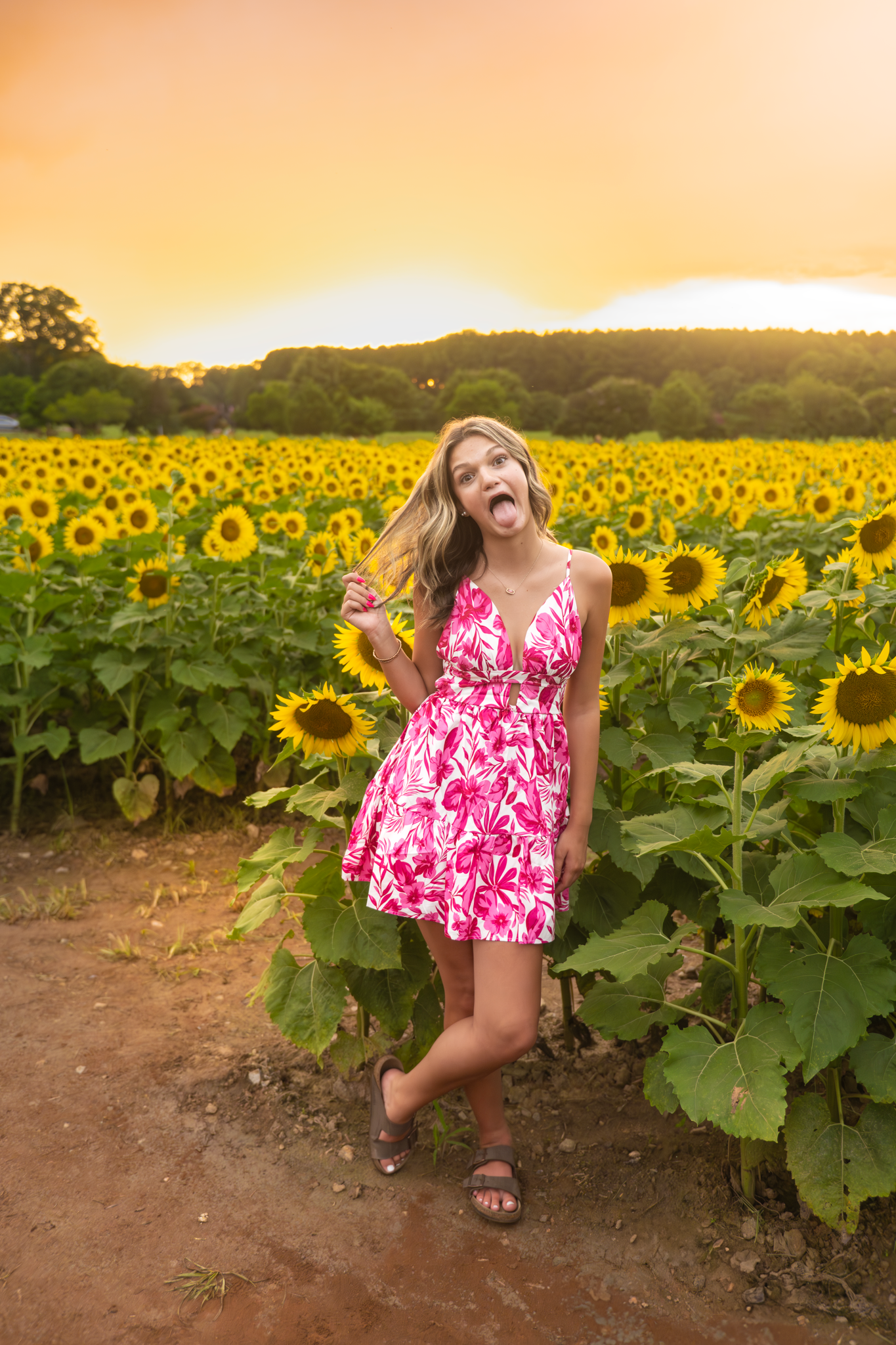 A woman in a pink floral dress poses playfully in a vibrant field of blooming sunflowers at sunset.