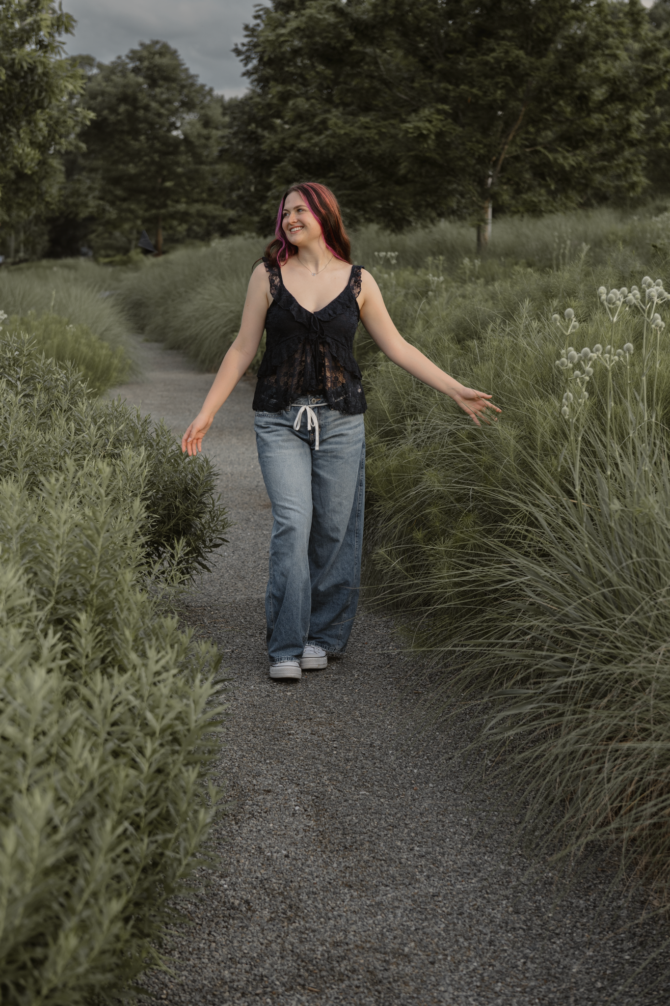Haylee walking along a winding path through tall grasses and trees on a cloudy day during her senior session in Raleigh, NC