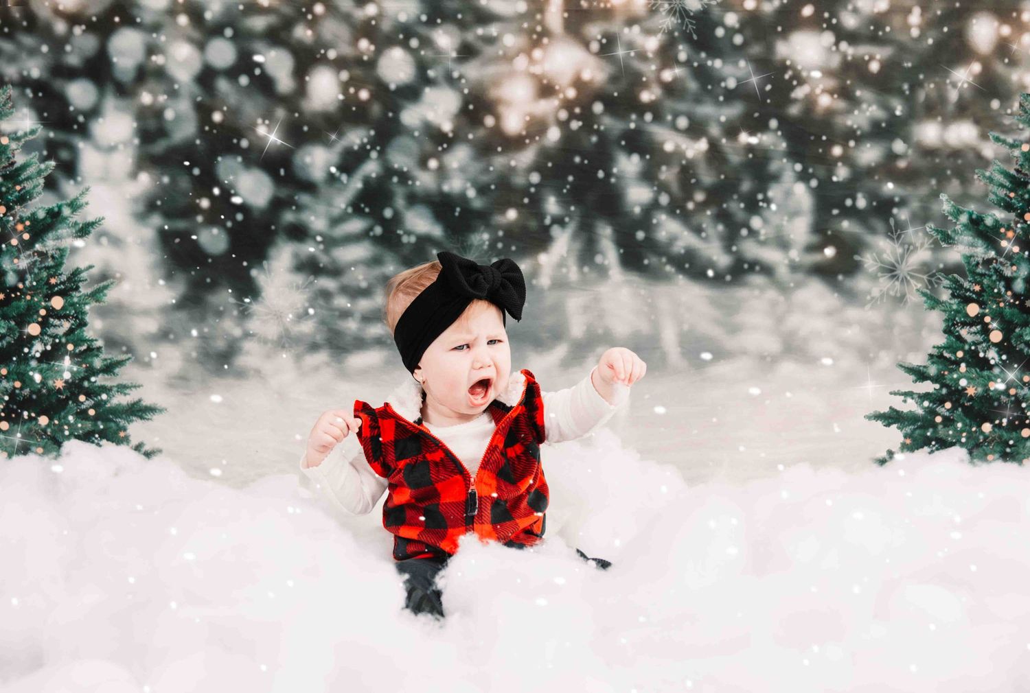 Small child in red buffalo plaid plays in the snow with evergreen trees and twinkling lights in background.