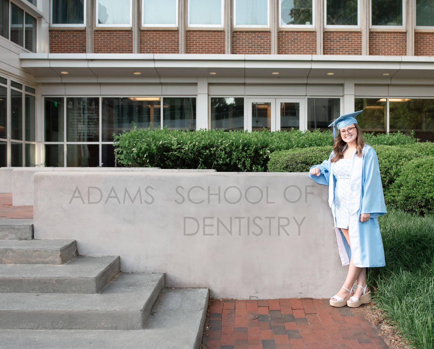 A graduate in blue cap and gown stands on the steps outside Adams School of Dentistry building.