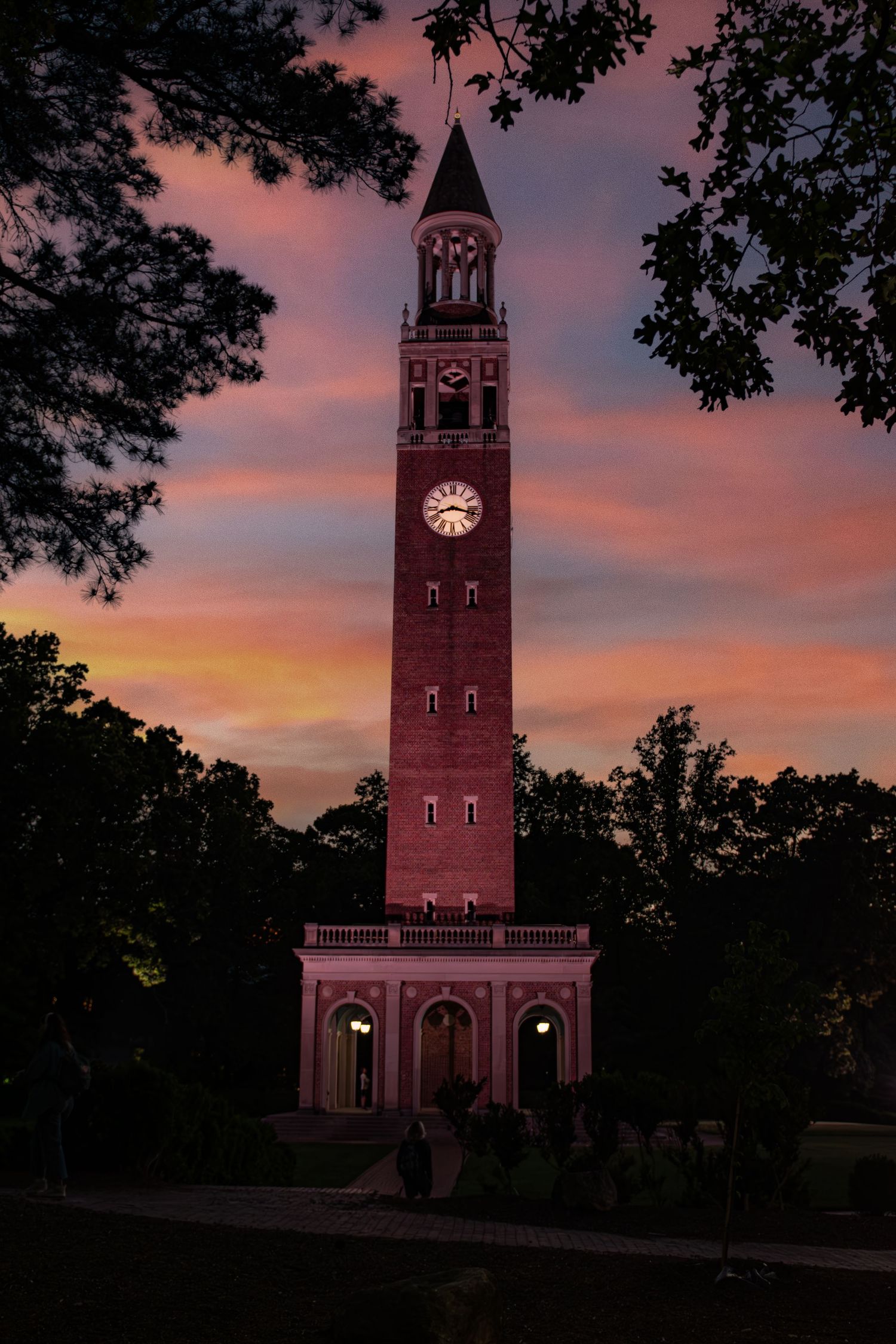 The iconic Morehead-Patterson Bell Tower at UNC Chapel Hill stands tall against a vibrant orange and purple sunset sky.