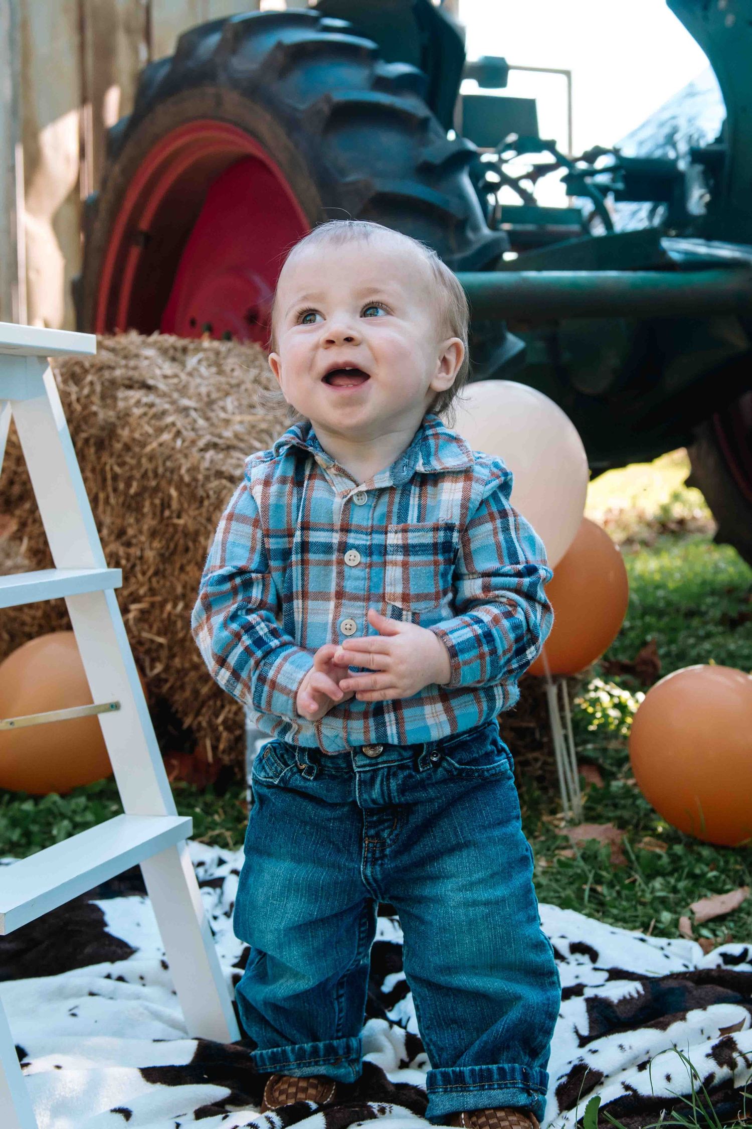 A young child in a plaid shirt and jeans stands smiling by hay bales and pumpkins with a tractor visible in the background.
