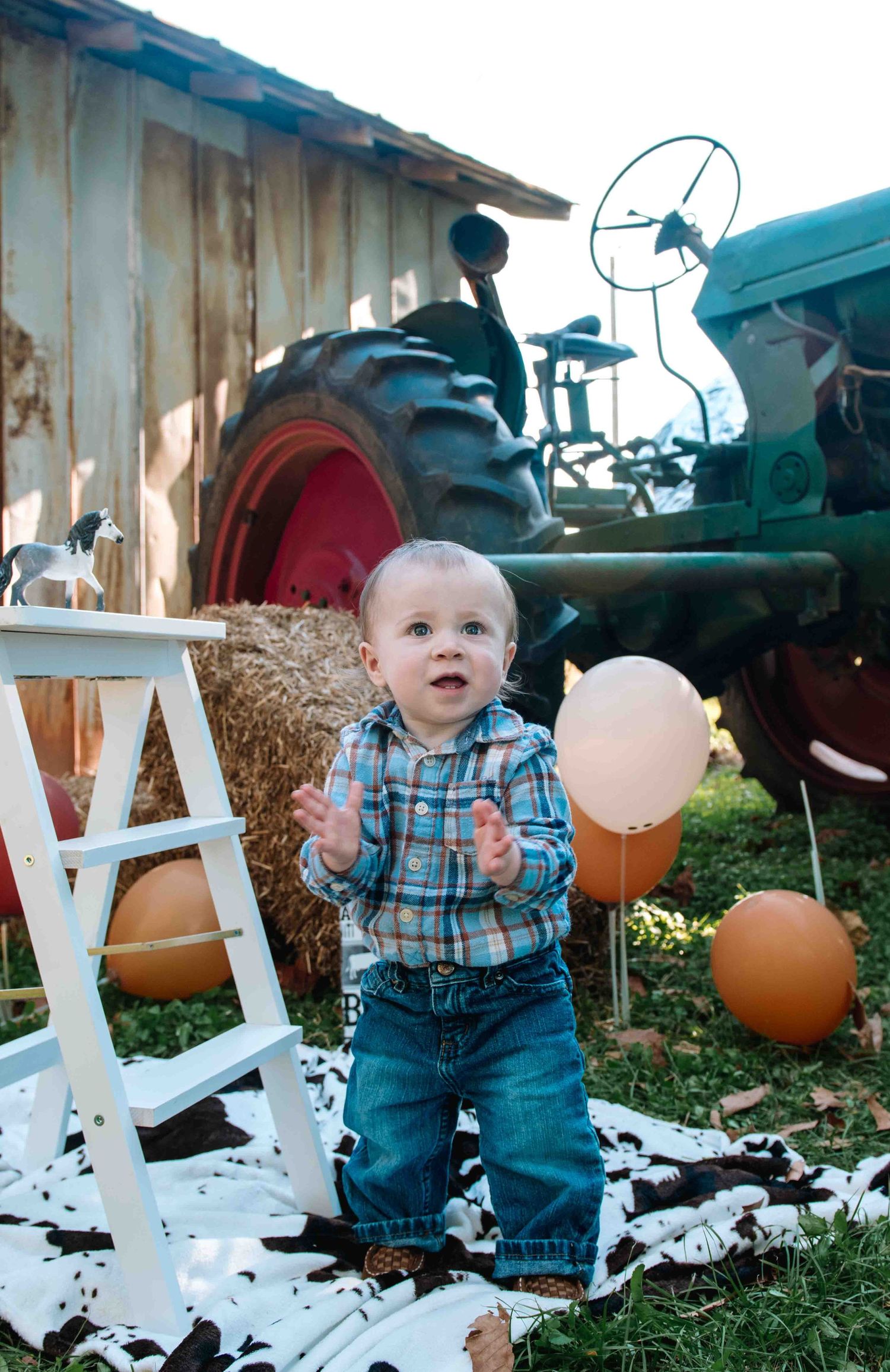 Small child in plaid shirt and jeans stands by vintage tractor with orange balloons and hay bales on farm.