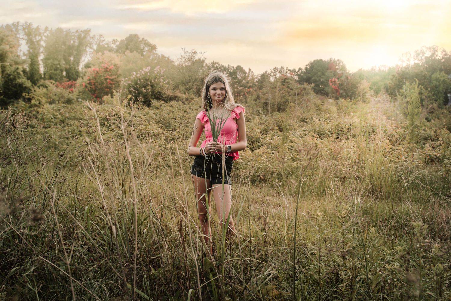 A person in a pink top stands in a golden sunlit field of tall grass during a summer evening.