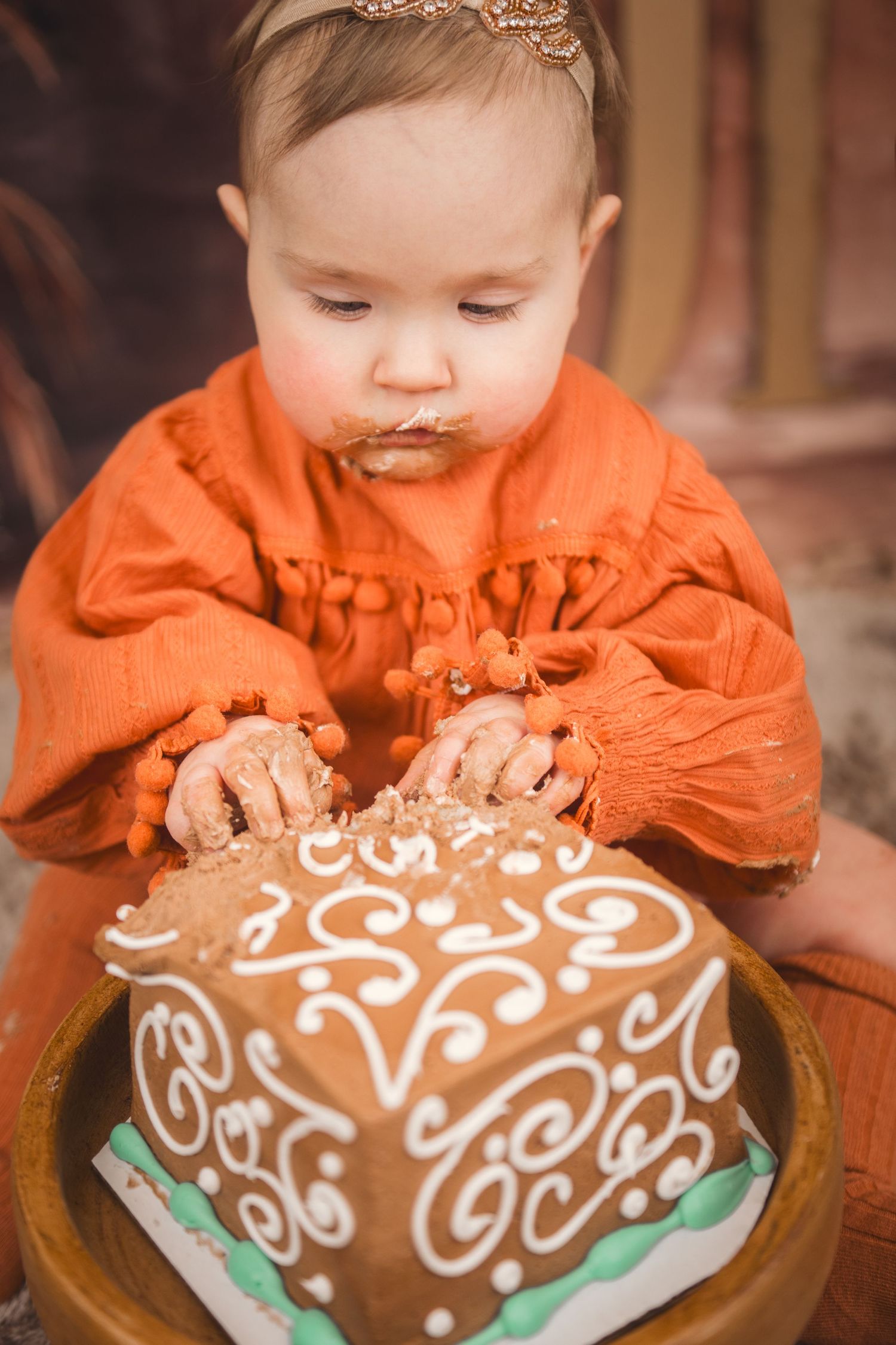 A small child in an orange dress explores a decorated chocolate cake with white frosting designs.