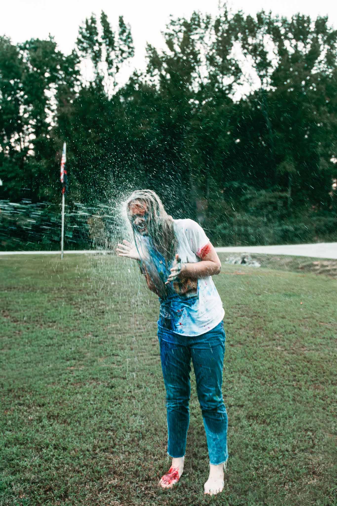 Person in casual clothes gets sprayed with water or liquid while jumping on a grassy field.