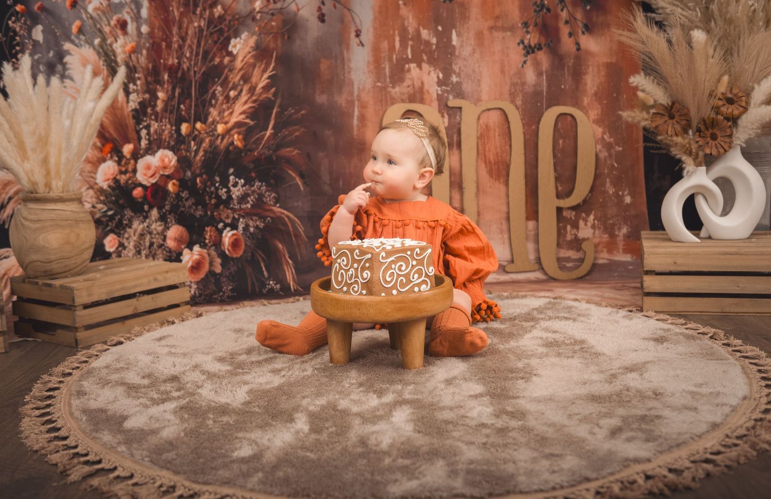 Child in orange clothing sits with pumpkin on circular rug surrounded by fall decor and dried floral arrangements.