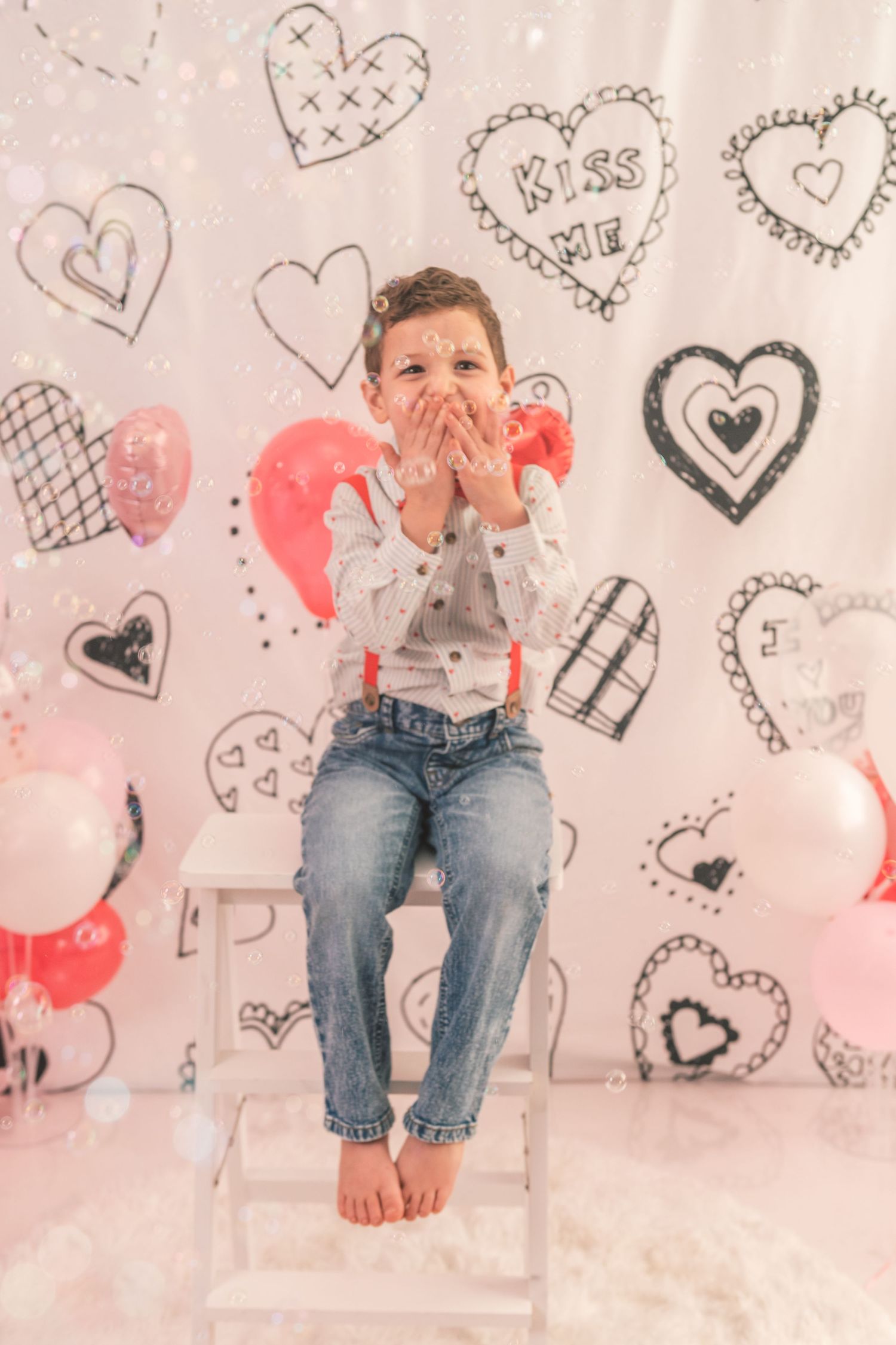 Cute Valentine's Day themed photo setup with hand drawn hearts on wall, red balloons and sitting pose against white backdrop.