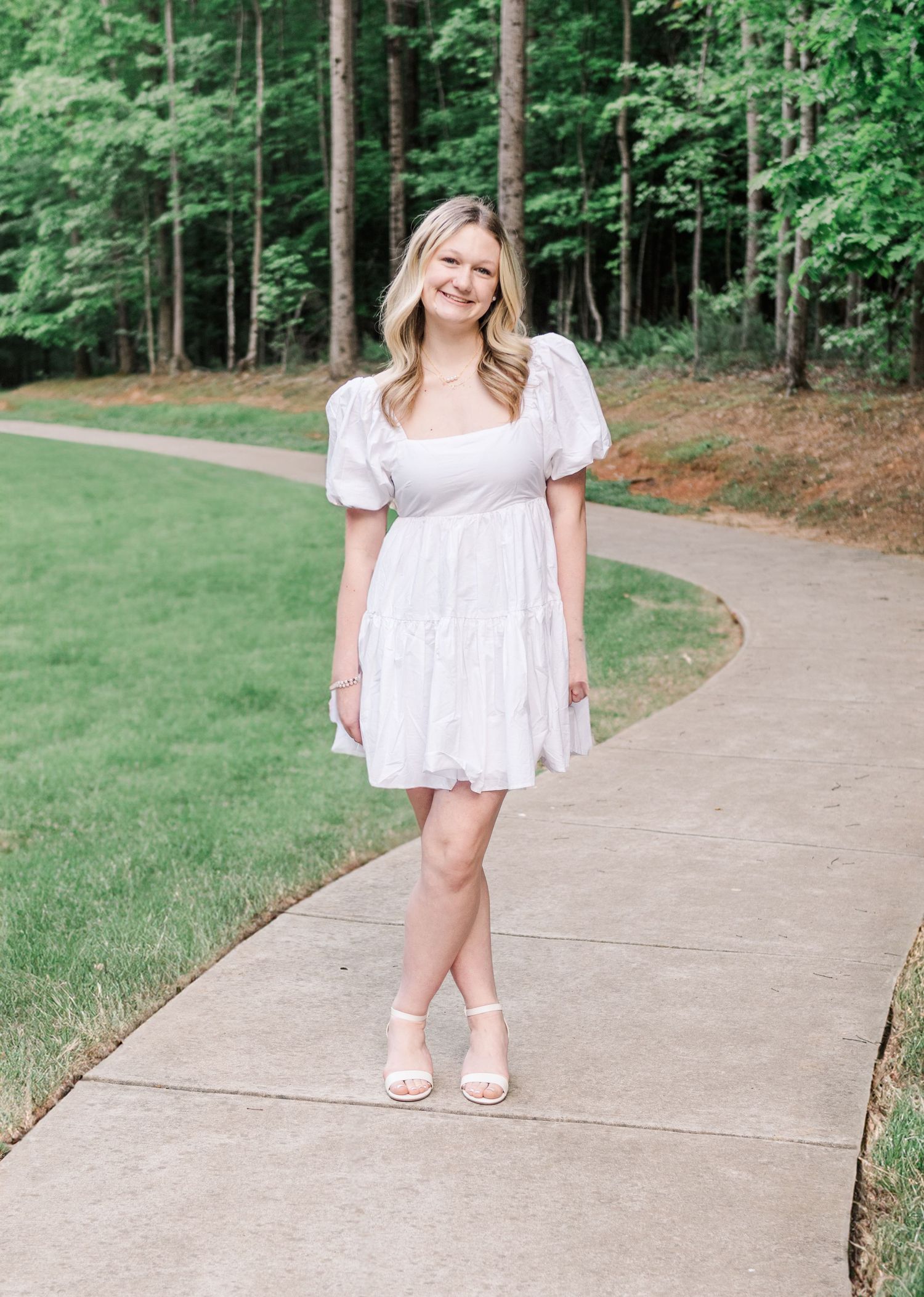 Person in a white eyelet summer dress with puff sleeves stands on a curved walkway surrounded by green trees and lawn.