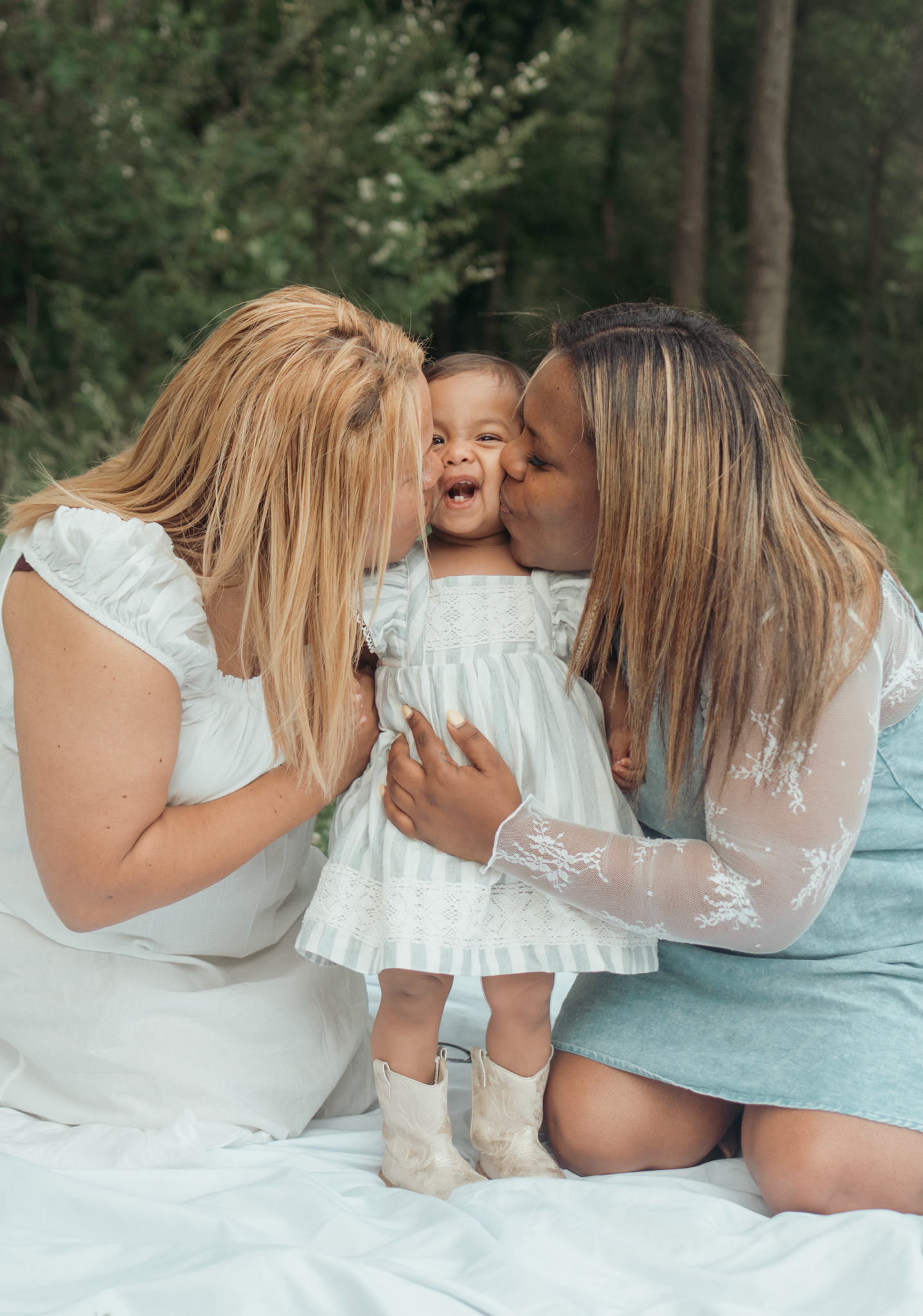 A joyful family moment captured outdoors on a white blanket as they share kisses and cuddles together in matching white outfits.