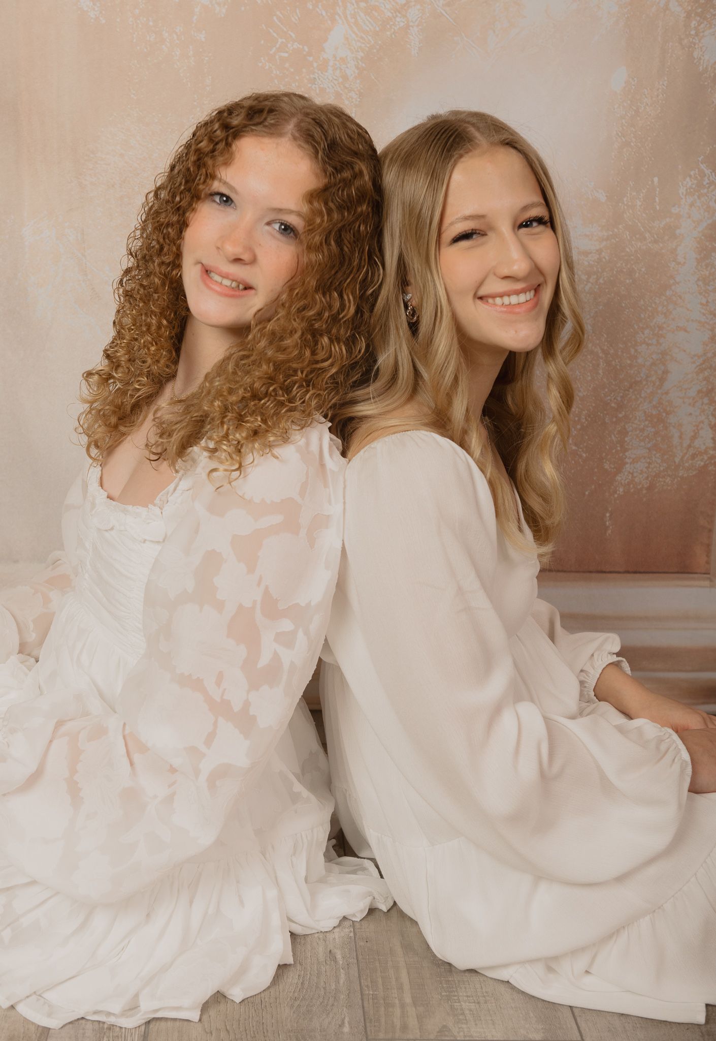 Two women wearing white dresses sit back-to-back, one with curly hair and one with straight blonde hair against beige wall.