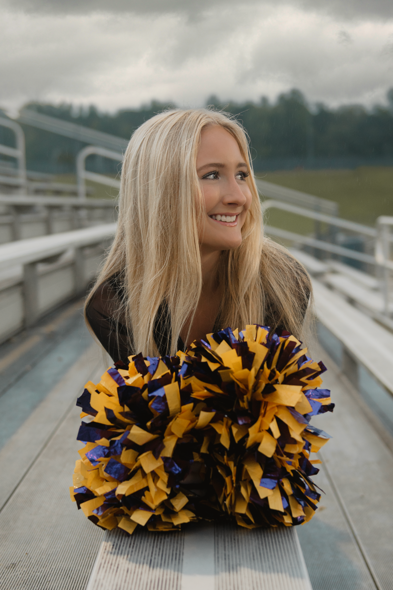 Cheerleader with long blonde hair smiles while holding blue and gold pom poms on metal bleachers.
