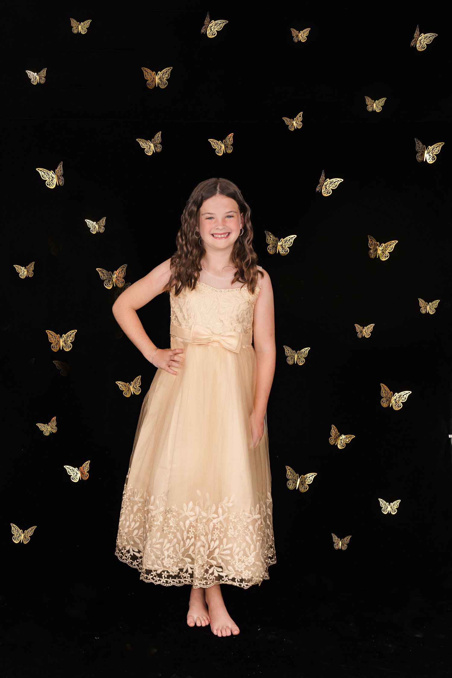 A barefoot subject in an elegant cream-colored dress poses against a black backdrop with golden butterfly decorations.