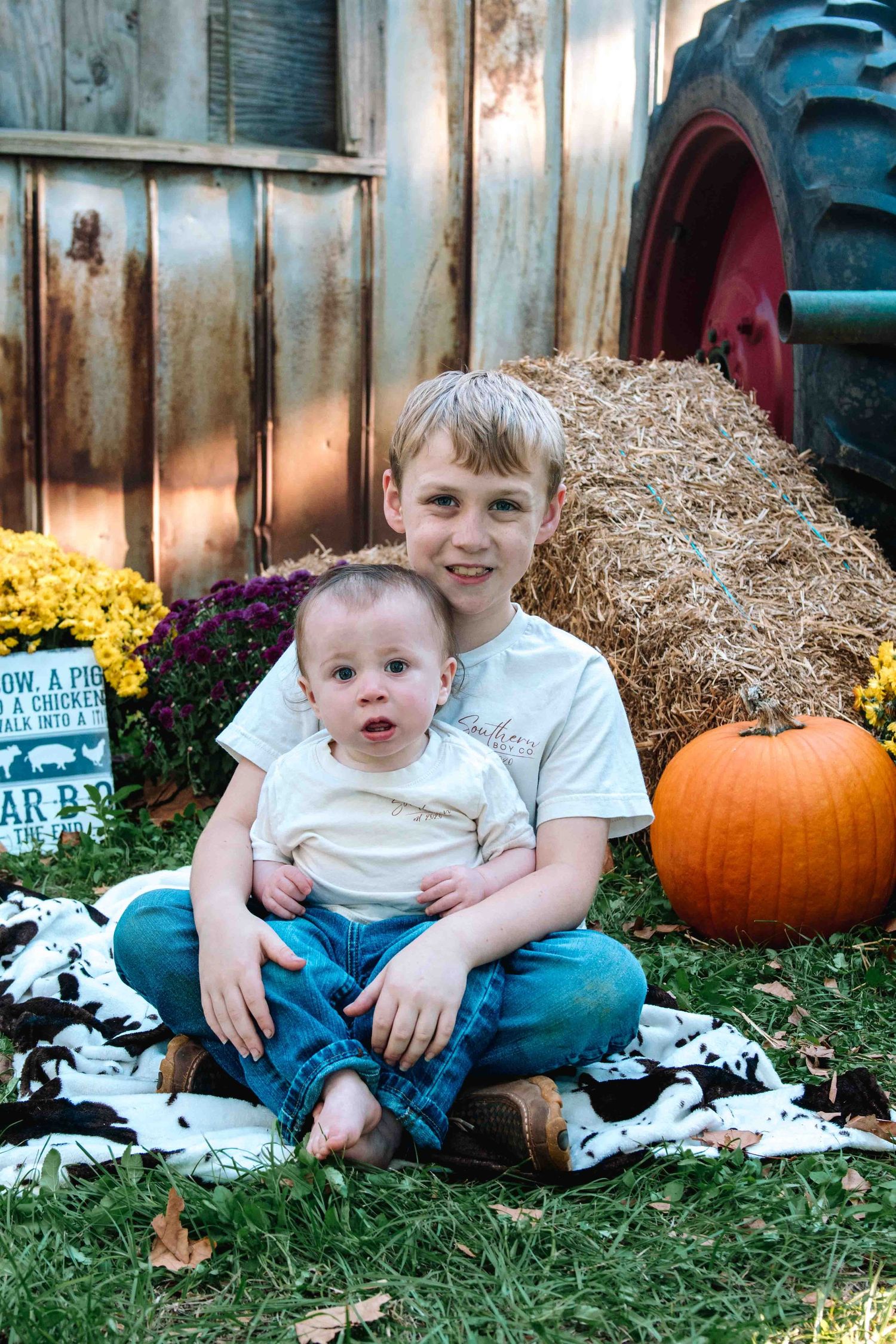 Autumn scene with pumpkin, mums and hay bale arranged with two young figures sitting on blanket outdoors.