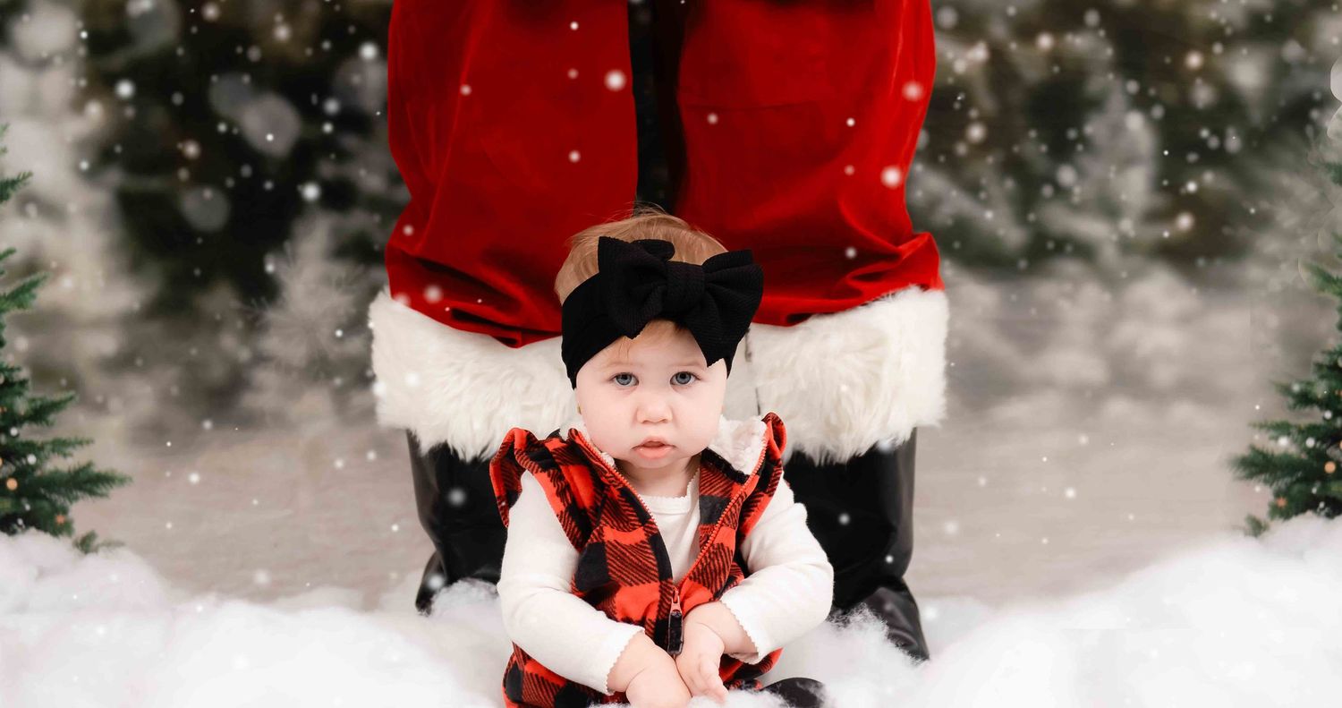A baby wearing red buffalo plaid and a black beanie poses in front of Santa's legs in a snowy setting.