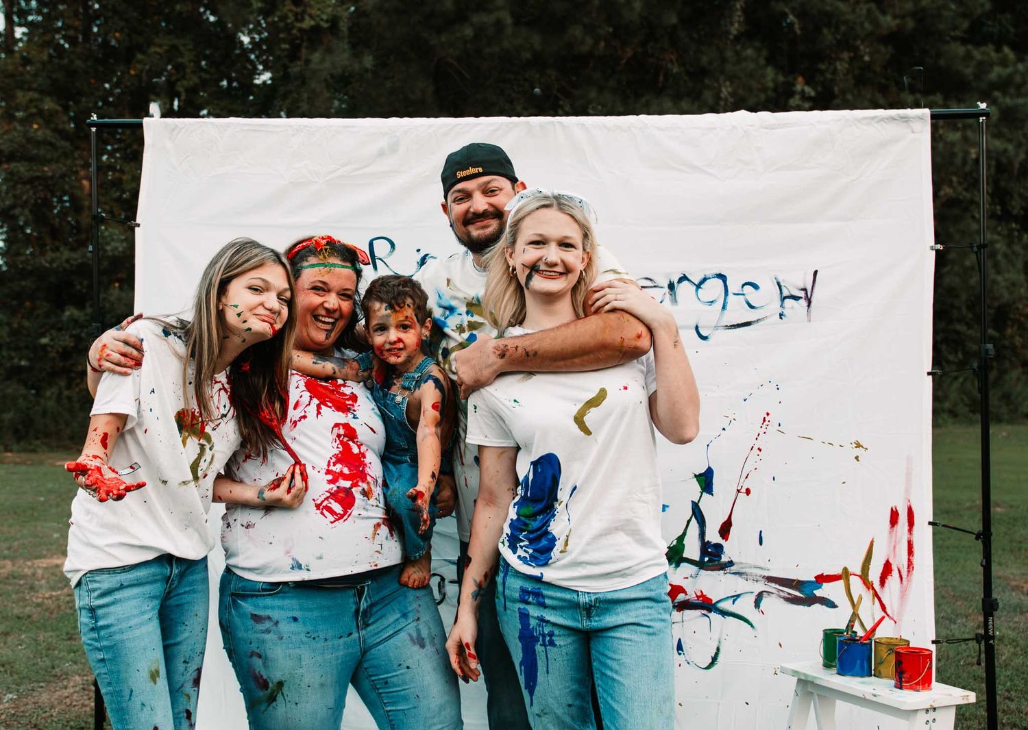 A group wearing white t-shirts and jeans poses together in front of a paint-splattered backdrop outdoors.