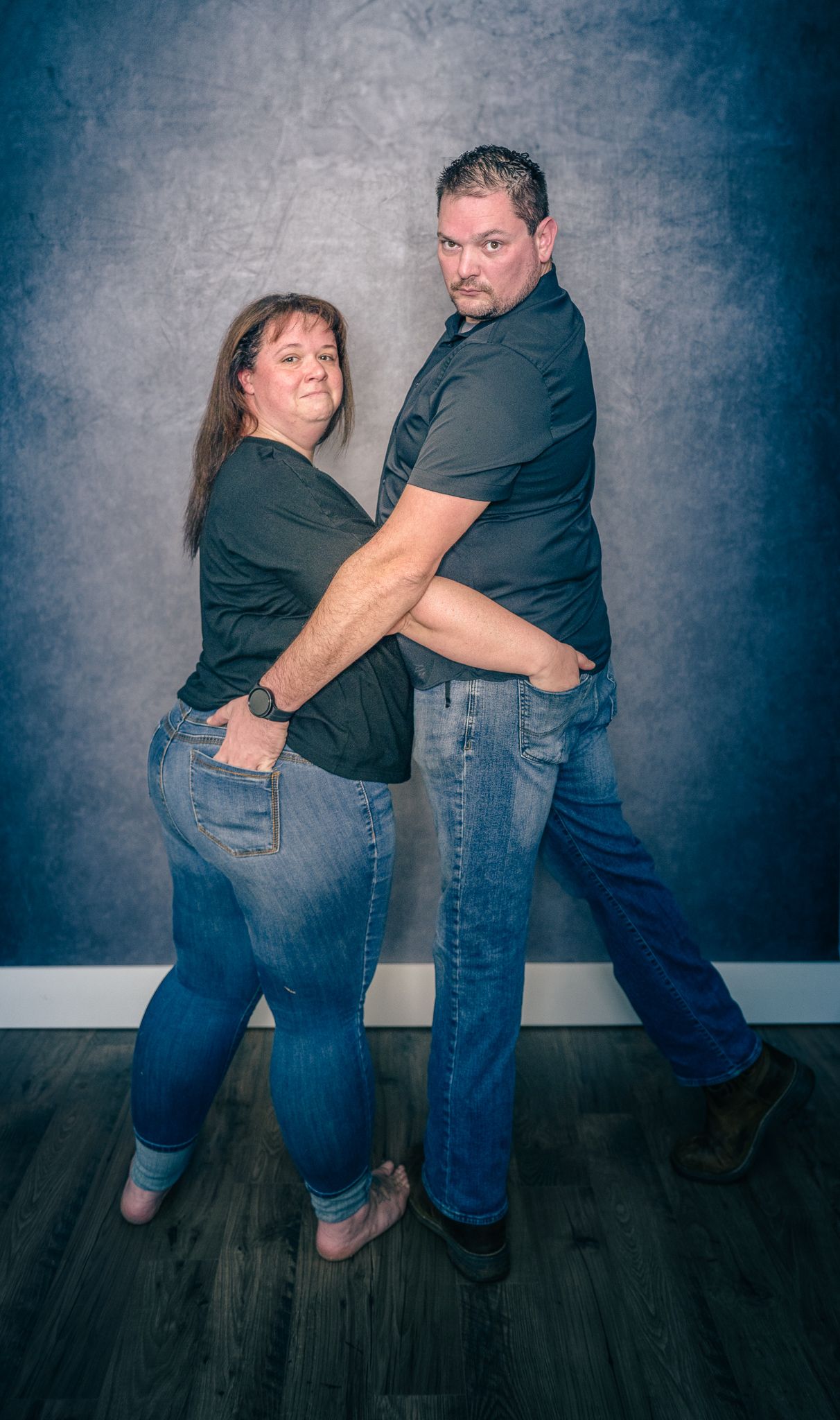 A couple in casual wear poses playfully together on a dark wood floor against a blue-gray backdrop in a studio setting.
